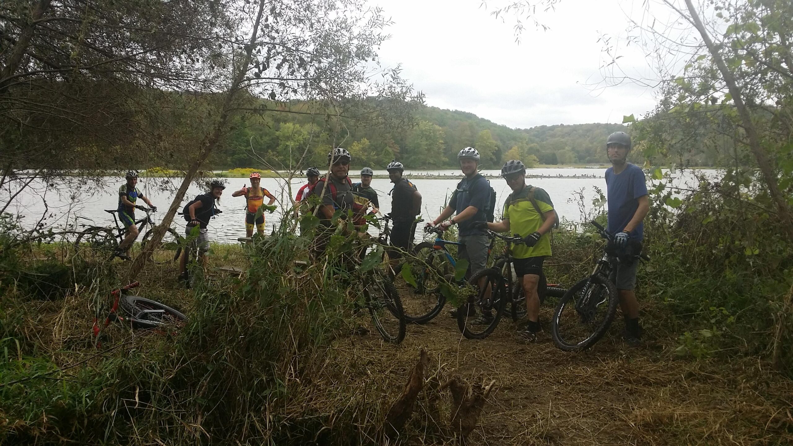 A group of mountain bikers gathered near a lake, with several bicycles resting on the ground. The scene features a mixture of greenery and water, set against a backdrop of rolling hills. The cyclists are casually interacting, some positioned next to their bikes while others stand by the water. It appears to be a cloudy day, suggesting an outdoor adventure in nature. Versailles State Park mountain bike trail.