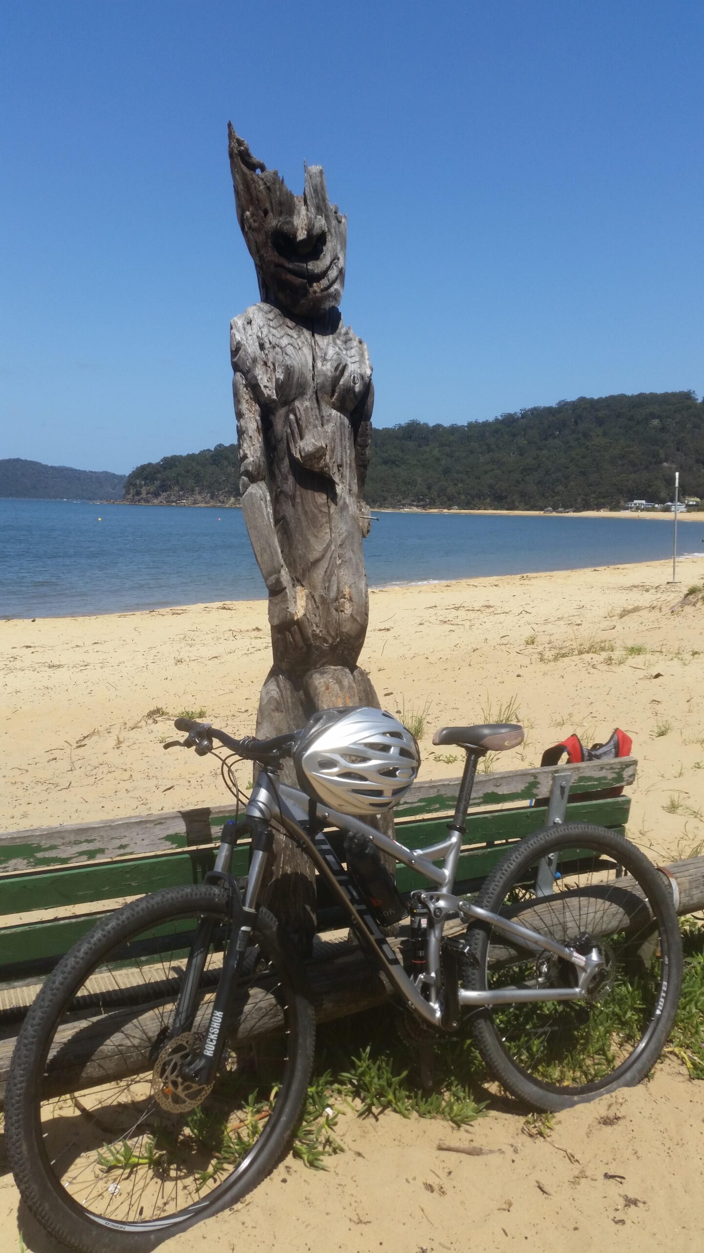 Norco Fluid 7.1: A wooden sculpture resembling a figure stands on the beach, with a clear blue sky above and a lush green hill in the background. In the foreground, a mountain bike with a silver helmet rests against a wooden bench on the sandy shore.