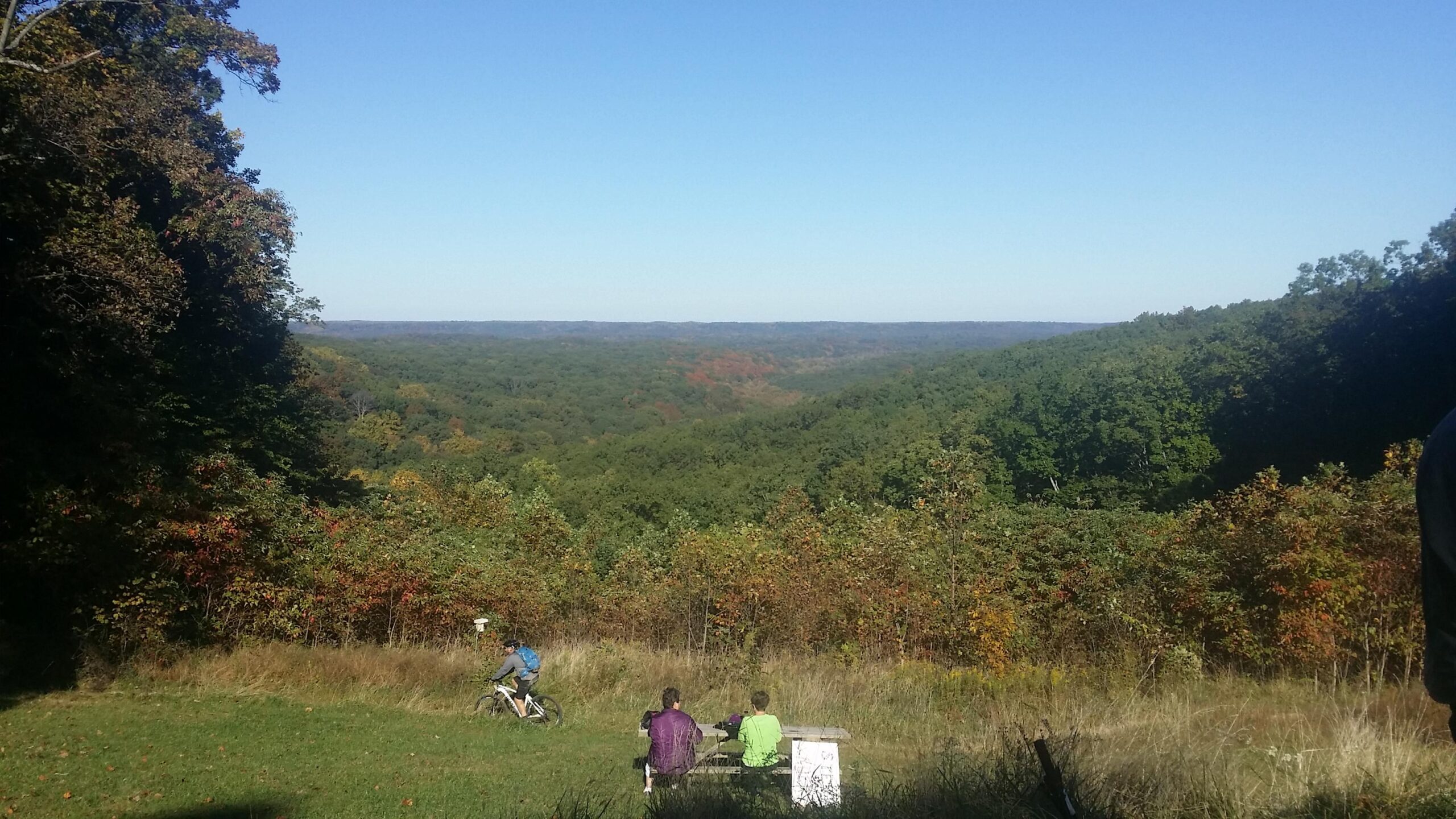 A scenic landscape view showcasing rolling hills covered in green trees with hints of autumn colors. In the foreground, two people sit at a picnic table, gazing at the vista, while a cyclist rides along a grassy path. The sky is clear and blue, adding to the tranquil atmosphere of the outdoor setting. Brown County Park mountain bike trail.