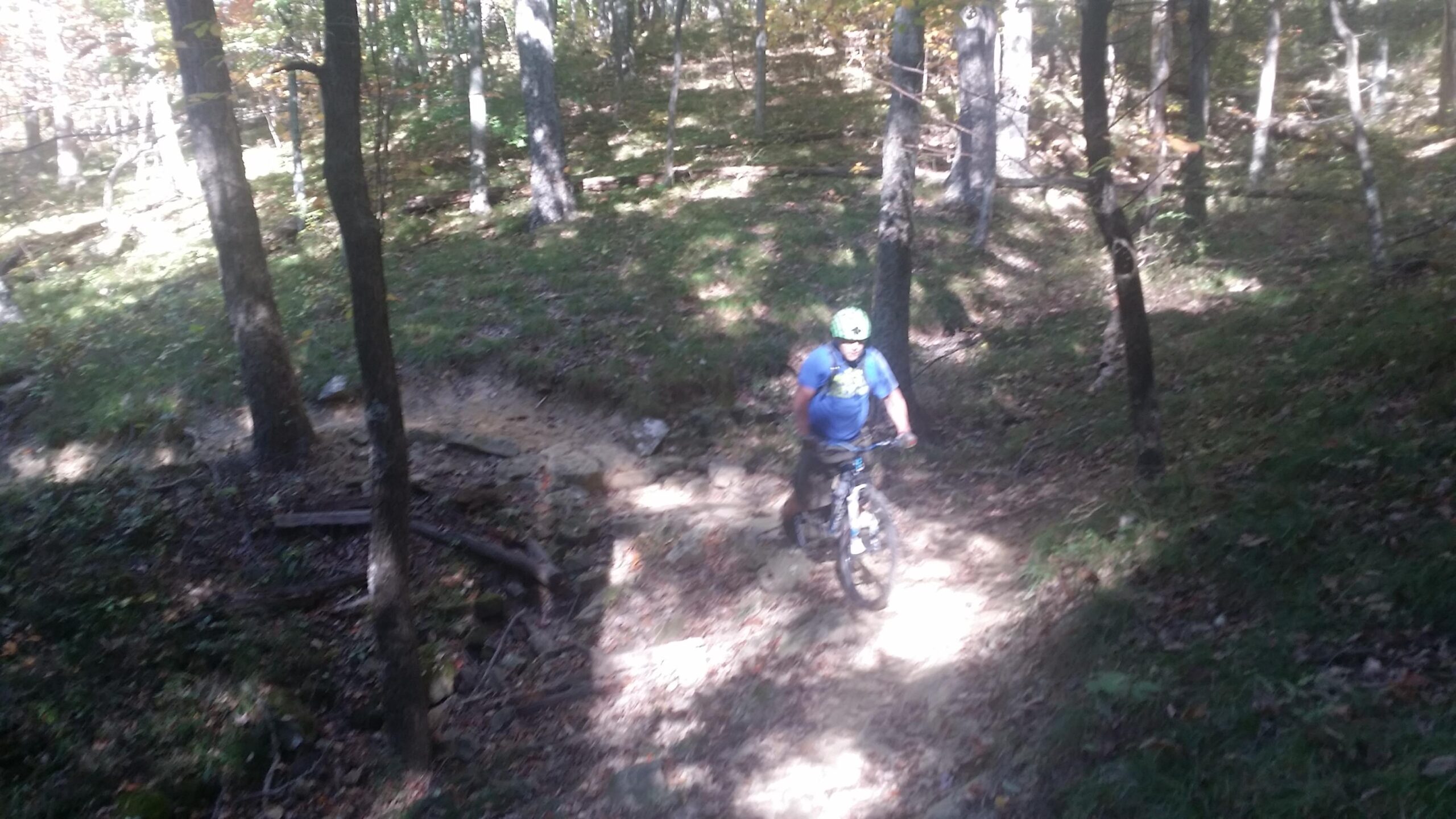 A mountain biker riding on a dirt trail surrounded by trees in a wooded area. The sunlight filters through the foliage, creating a mix of light and shadow on the ground. Brown County Park mountain bike trail.