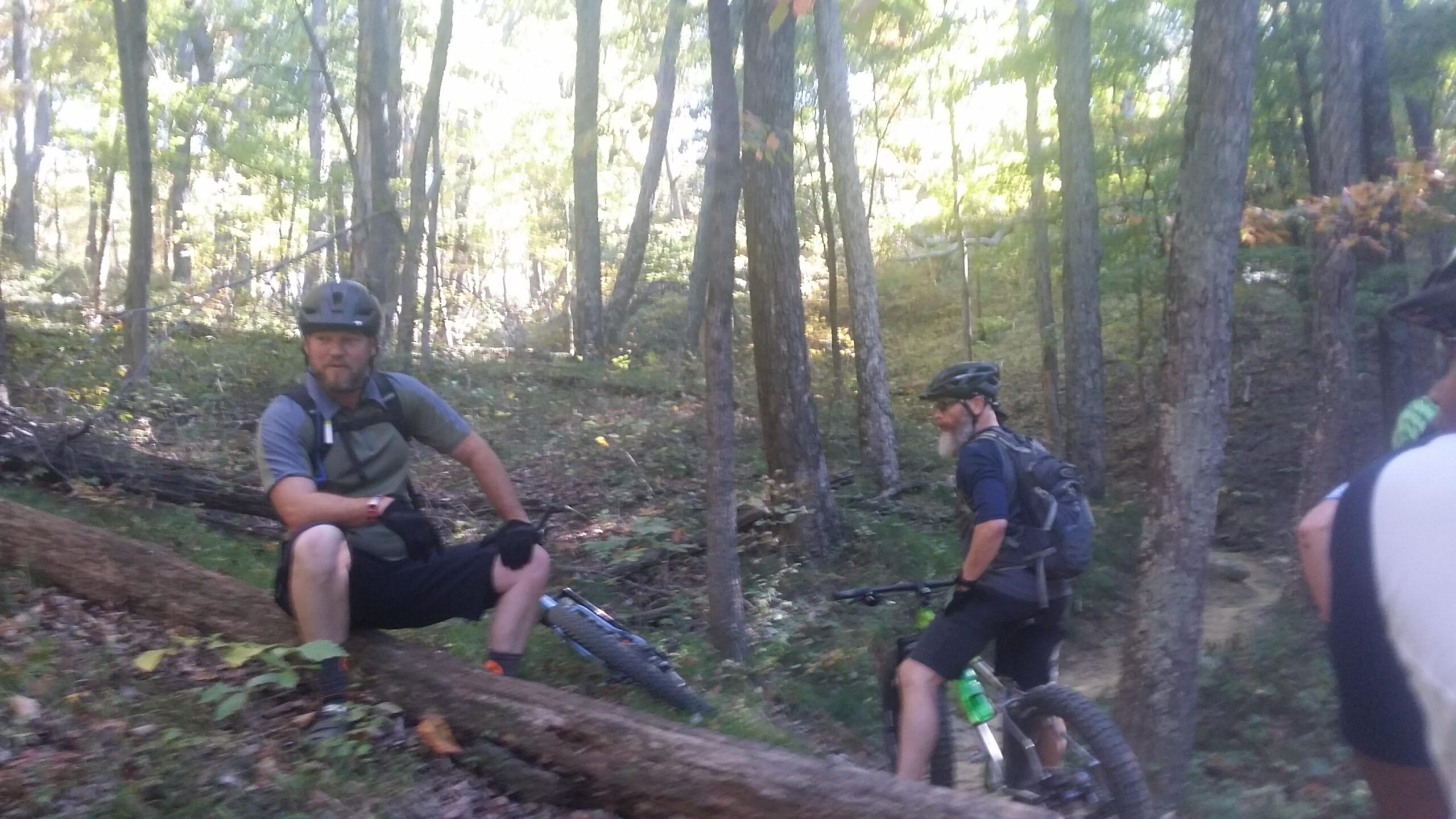 Two mountain bikers are resting on a trail in a wooded area. One biker is sitting on a fallen log, wearing a helmet and casual biking attire. The second biker stands nearby, also wearing a helmet and equipped with a backpack. The background features tall trees and lush greenery, indicating a serene outdoor environment. Brown County Park mountain bike trail.