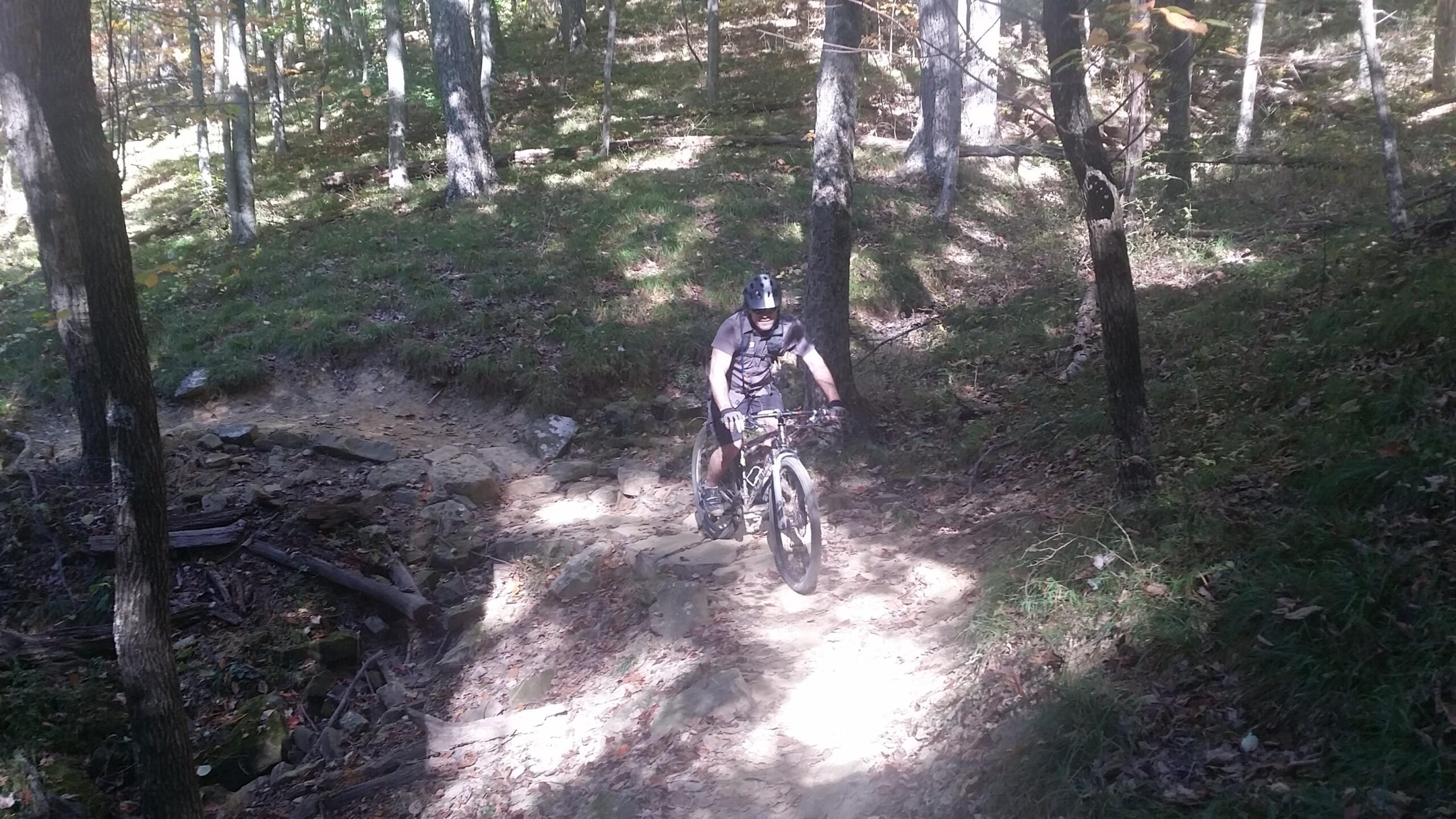 A mountain biker navigating a rocky dirt trail in a forested area, surrounded by trees and patches of sunlight filtering through the canopy. Brown County Park mountain bike trail.