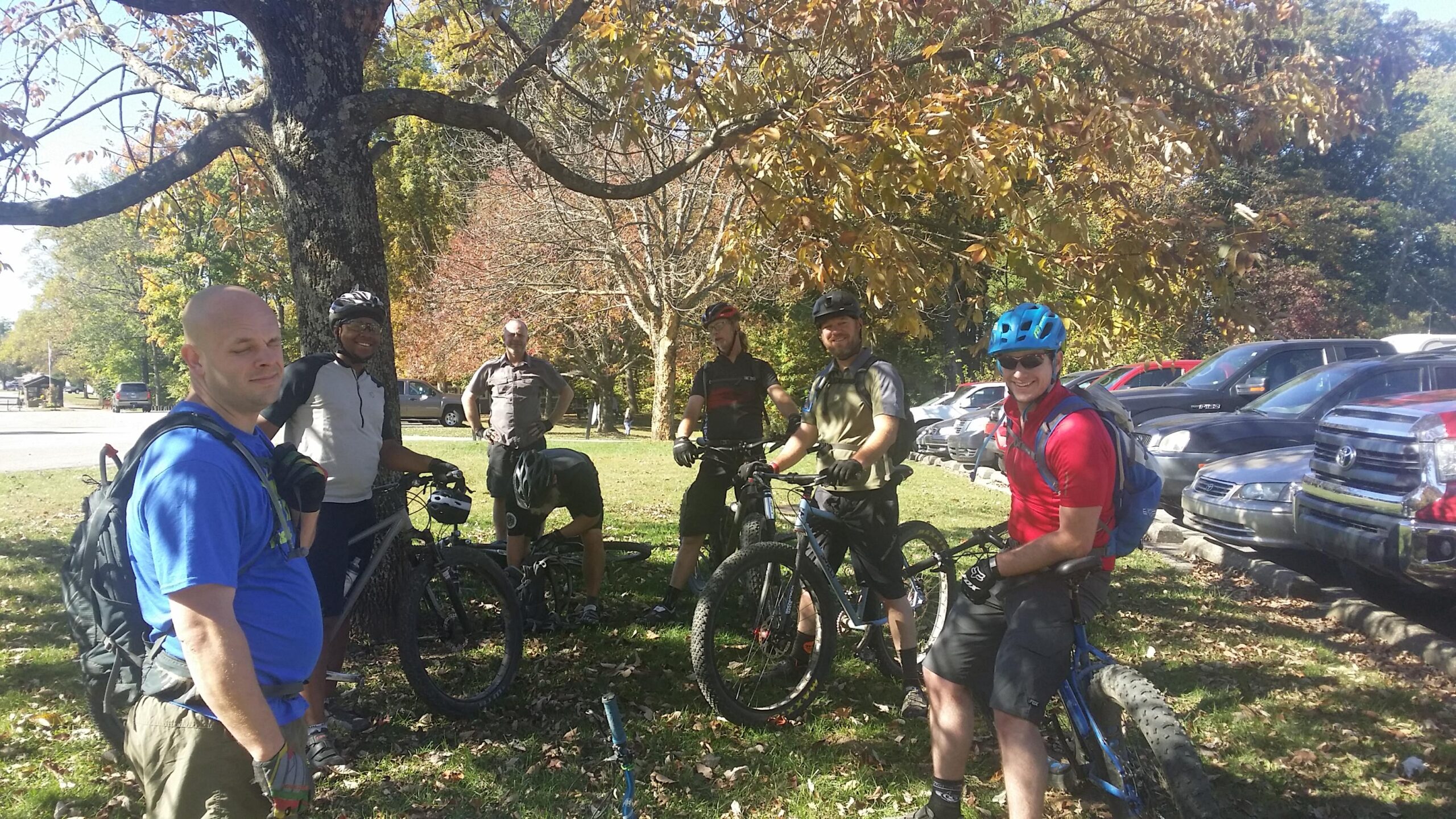 Group of mountain bikers gathered under a tree in a parking area, dressed in cycling gear and helmets. It’s a sunny day with colorful autumn leaves in the background, and several parked vehicles are visible. Some bikers are standing next to their bikes, while one is crouched down checking their equipment. Brown County Park mountain bike trail.