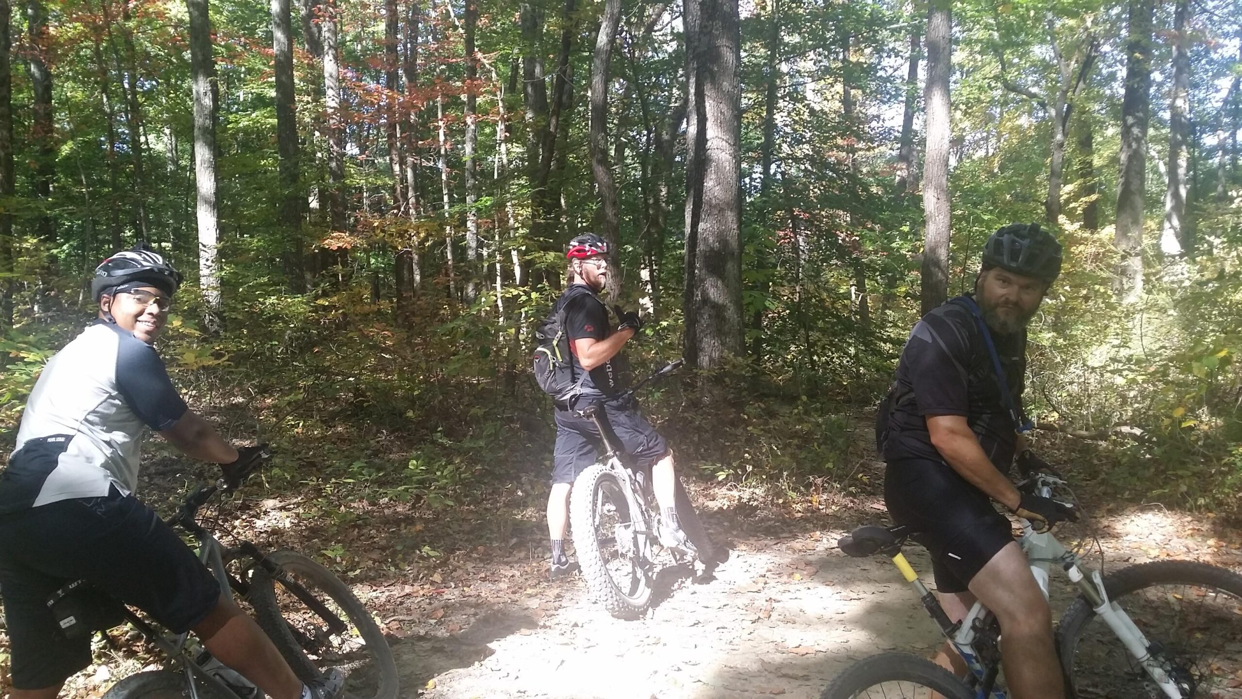 Three mountain bikers pause on a dirt trail surrounded by trees in a forest. The sunlight filters through the leaves, creating a peaceful outdoor atmosphere. The bikers are wearing helmets and cycling gear, and they appear to be enjoying their ride. One is smiling at the camera, while the others are looking back, engaged in conversation. The trail and foliage indicate a scenic nature setting, perfect for biking enthusiasts. Brown County Park mountain bike trail.