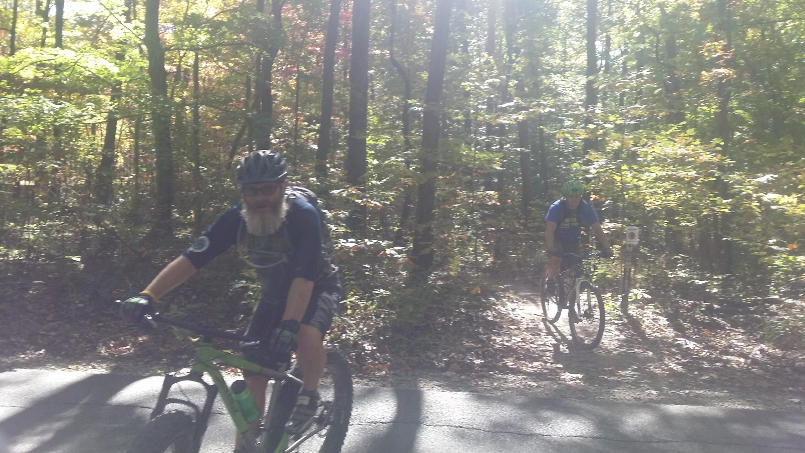 Two mountain bikers riding through a forested trail with sunlight filtering through the trees. The first biker, wearing a helmet and gloves, has a beard and is on a green bike, while the second biker in a blue shirt rides behind on a different bike. The surroundings are lush with greenery and hints of fall colors. Brown County Park mountain bike trail.