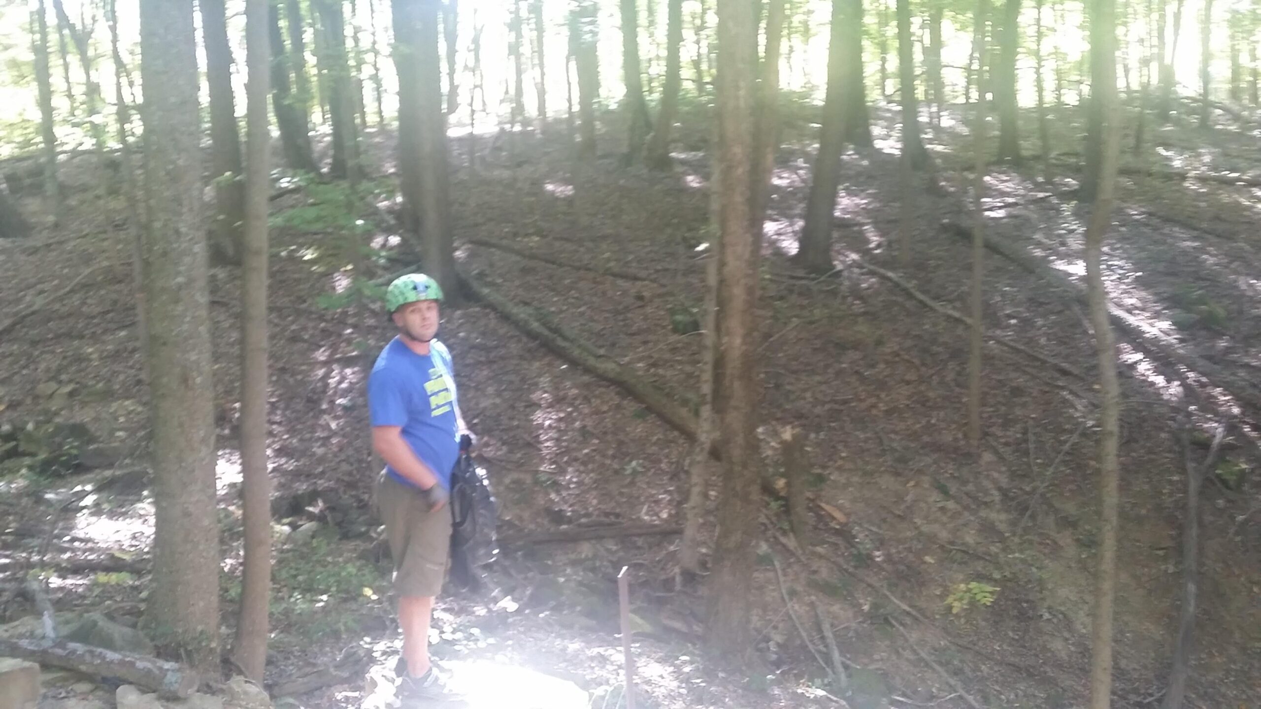 A person wearing a green helmet and a blue shirt stands in a wooded area, surrounded by trees and dappled sunlight filtering through the leaves. The ground is covered with fallen leaves and branches, creating a natural, earthy landscape. Brown County Park mountain bike trail.