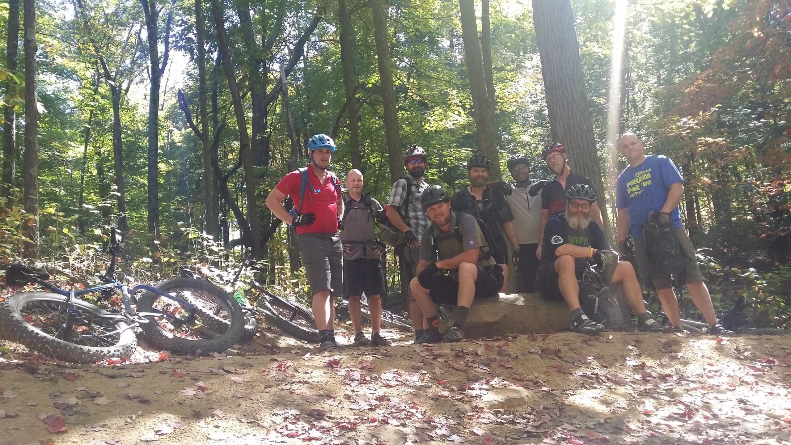 A group of nine mountain bikers posing for a photo on a dirt trail surrounded by trees. Several bikes are resting on the ground nearby, and the scene is brightly lit by sunlight filtering through the foliage. The riders are dressed in casual cycling attire and wearing helmets. Some are standing while others are sitting on a rock. Leaves are scattered on the ground, suggesting an autumn setting. Brown County Park mountain bike trail.