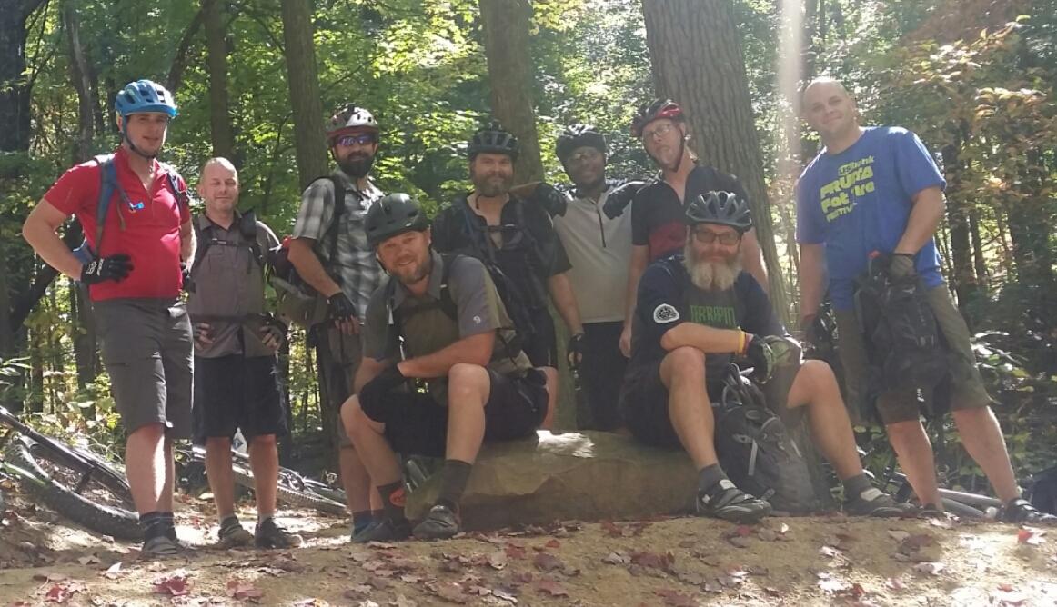 A group of nine men, wearing mountain biking helmets and gear, pose for a photo on a rock in a forested area. The sunlight filters through the trees, creating a bright and inviting atmosphere. Some men are smiling or making fun gestures, while others have a relaxed stance. Bicycles can be seen in the background, indicating they have been biking in the woods. Brown County Park mountain bike trail.