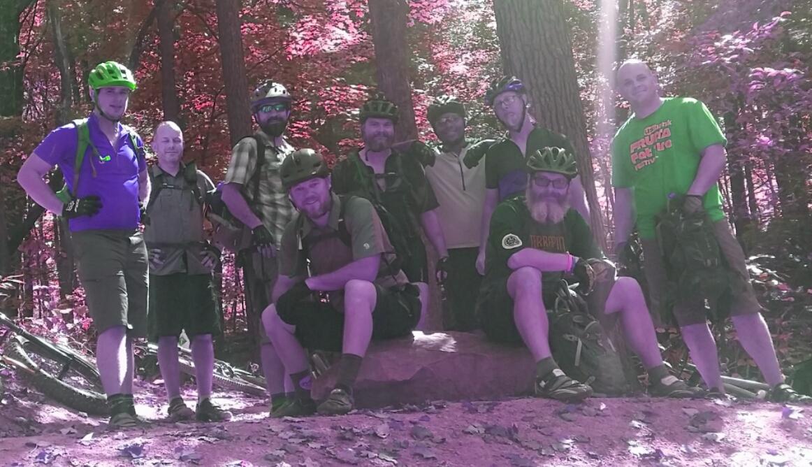 A group of nine men wearing biking gear and helmets pose together in a forested area, surrounded by trees with vibrant autumn foliage. They stand and sit on a large rock, smiling and engaging with the camera, with mountain bikes visible in the background. The image has a purple hue, adding a stylized effect to the scene. Brown County Park mountain bike trail.