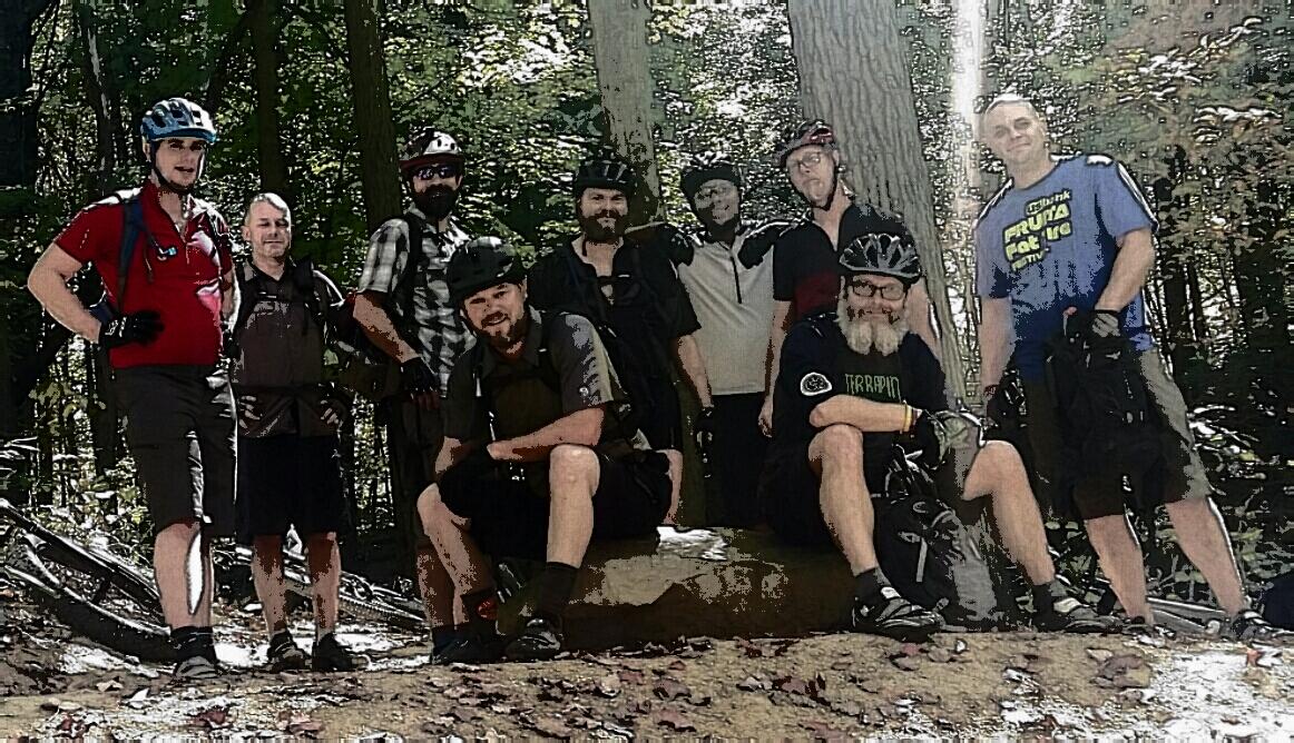 A group of nine men wearing cycling helmets and gear pose together in a forest setting. They are standing and sitting on a large rock, surrounded by trees and greenery, suggesting a mountain biking or outdoor adventure activity. The group appears friendly and relaxed, showcasing their love for biking and nature. Brown County Park mountain bike trail.