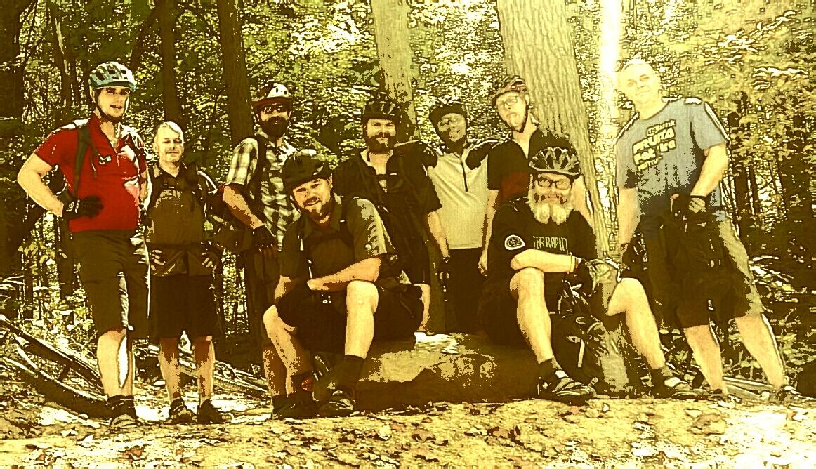A group of eight mountain bikers poses for a photo in a wooded area, wearing helmets and cycling gear. They are standing and sitting on a rock, surrounded by trees, with sunlight filtering through the leaves. The atmosphere is casual and friendly, showcasing their camaraderie during an outdoor biking adventure. Brown County Park mountain bike trail.