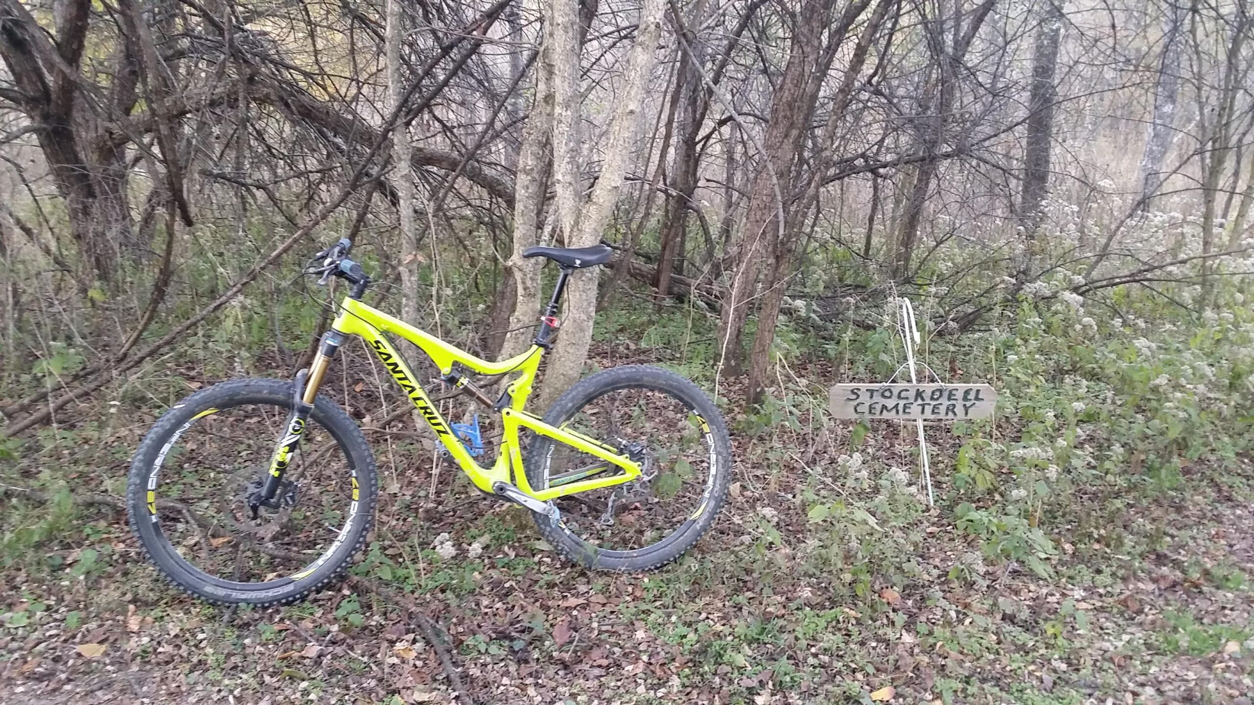 A vibrant yellow mountain bike is parked on a dirt path surrounded by dense trees and foliage. Nearby, a wooden sign reads "Stockdell Cemetery," partially obscured by the natural growth of plants and grass. The scene conveys a sense of adventure and exploration in a rustic outdoor setting. Skullbuster mountain bike trail.