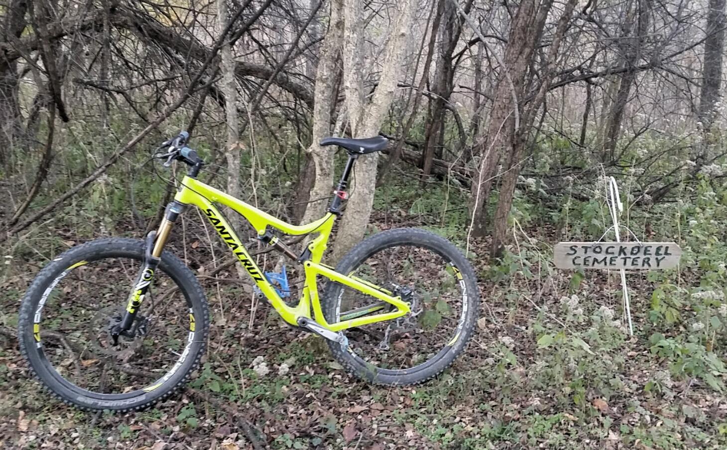 A bright yellow mountain bike is parked on the ground among dried leaves and green underbrush, next to a wooden sign that reads "Stock Dell Cemetery." The background features a dense cluster of trees and branches, creating a natural, wooded setting. Skullbuster mountain bike trail.