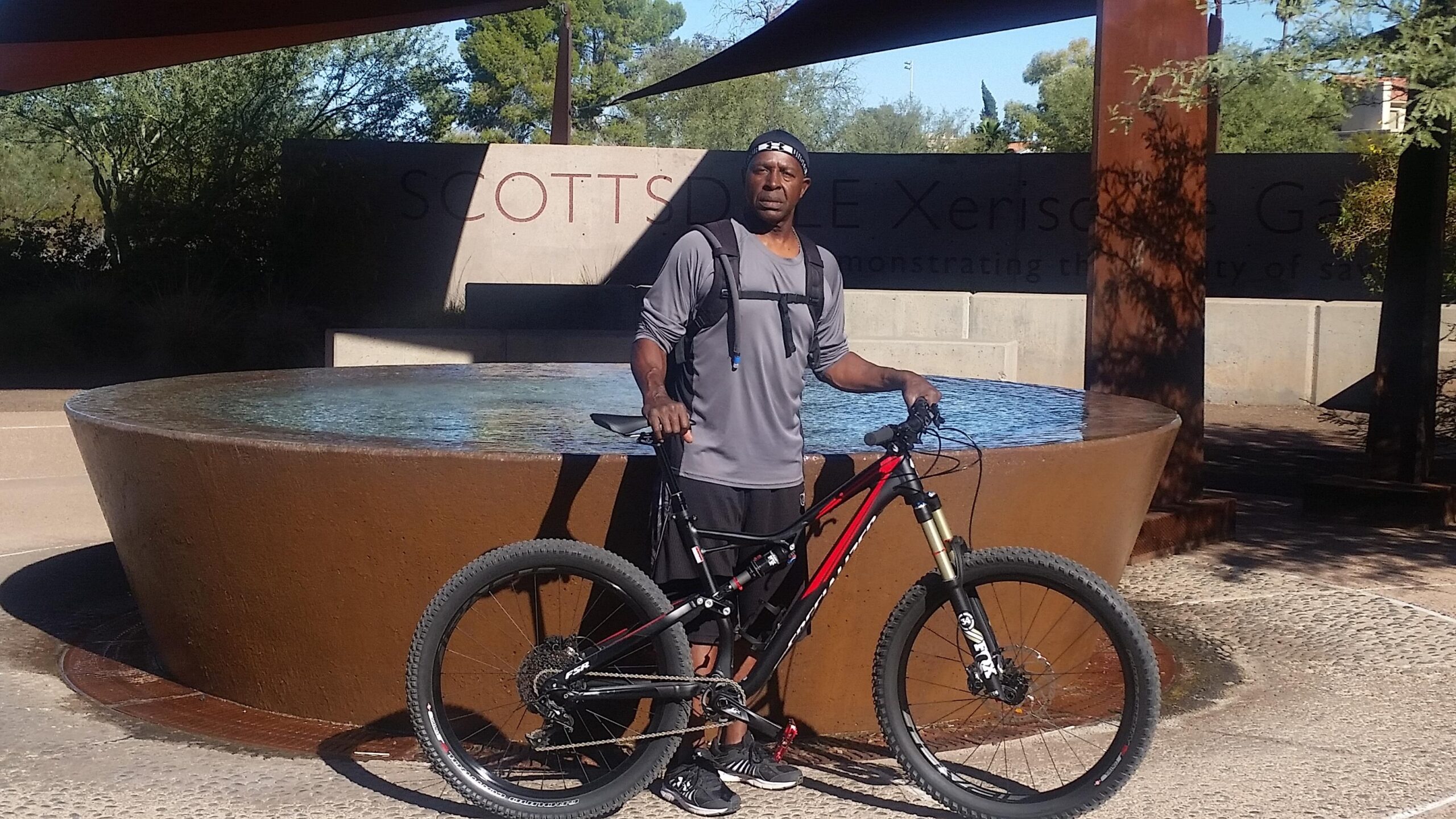 Specialized Stumpjumper FSR 6Fattie: A person stands next to a mountain bike, wearing athletic gear and a backpack. They are positioned in front of a large, round, water feature with a sign that reads "Scottsdale Xeriscape Garden" in the background, surrounded by greenery on a sunny day.