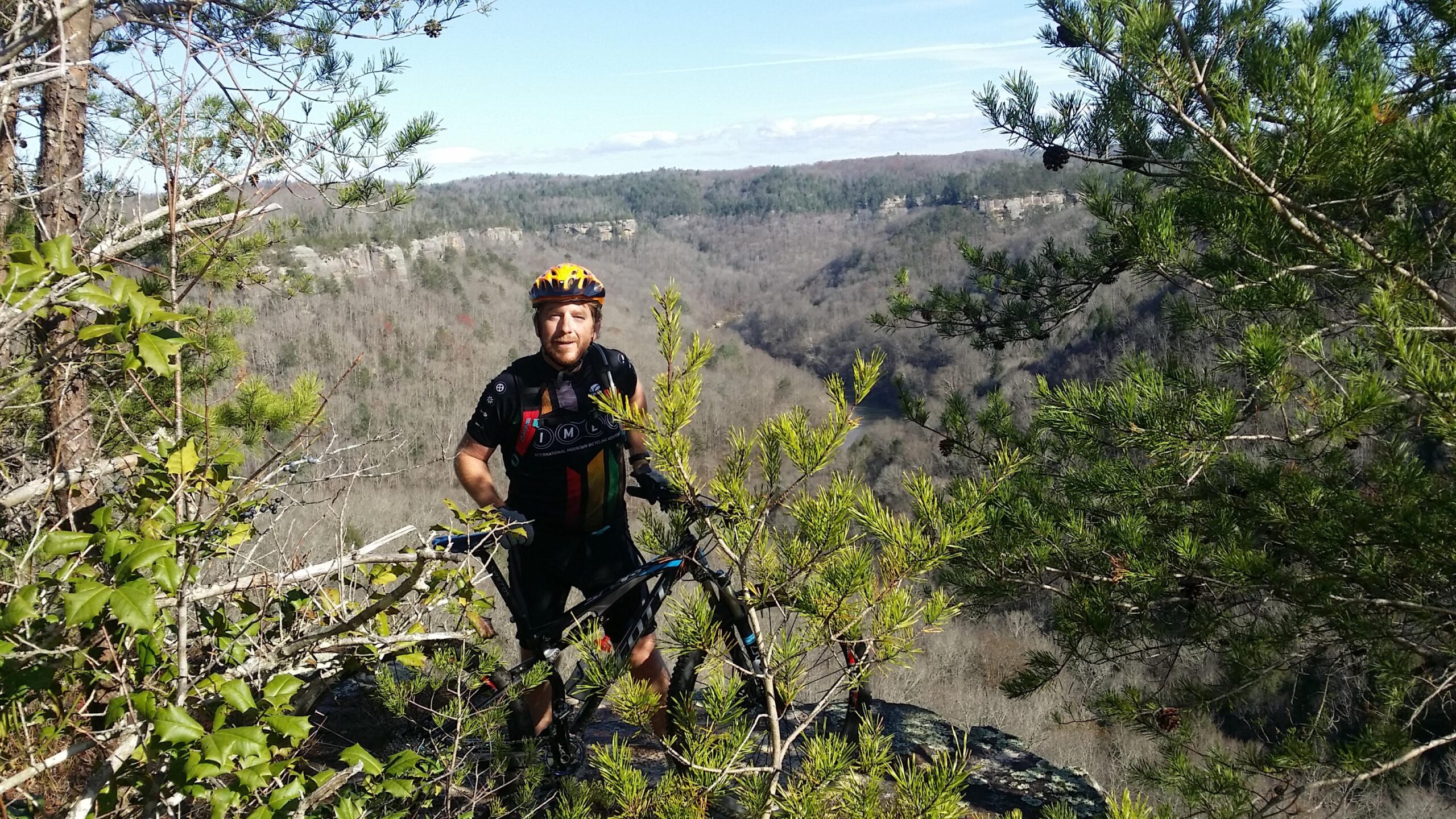 A person in cycling gear stands on a rocky outcrop surrounded by greenery, holding a mountain bike. The background features a scenic view of a valley with trees covering the hills, under a clear blue sky. Grand Gap mountain bike trail.