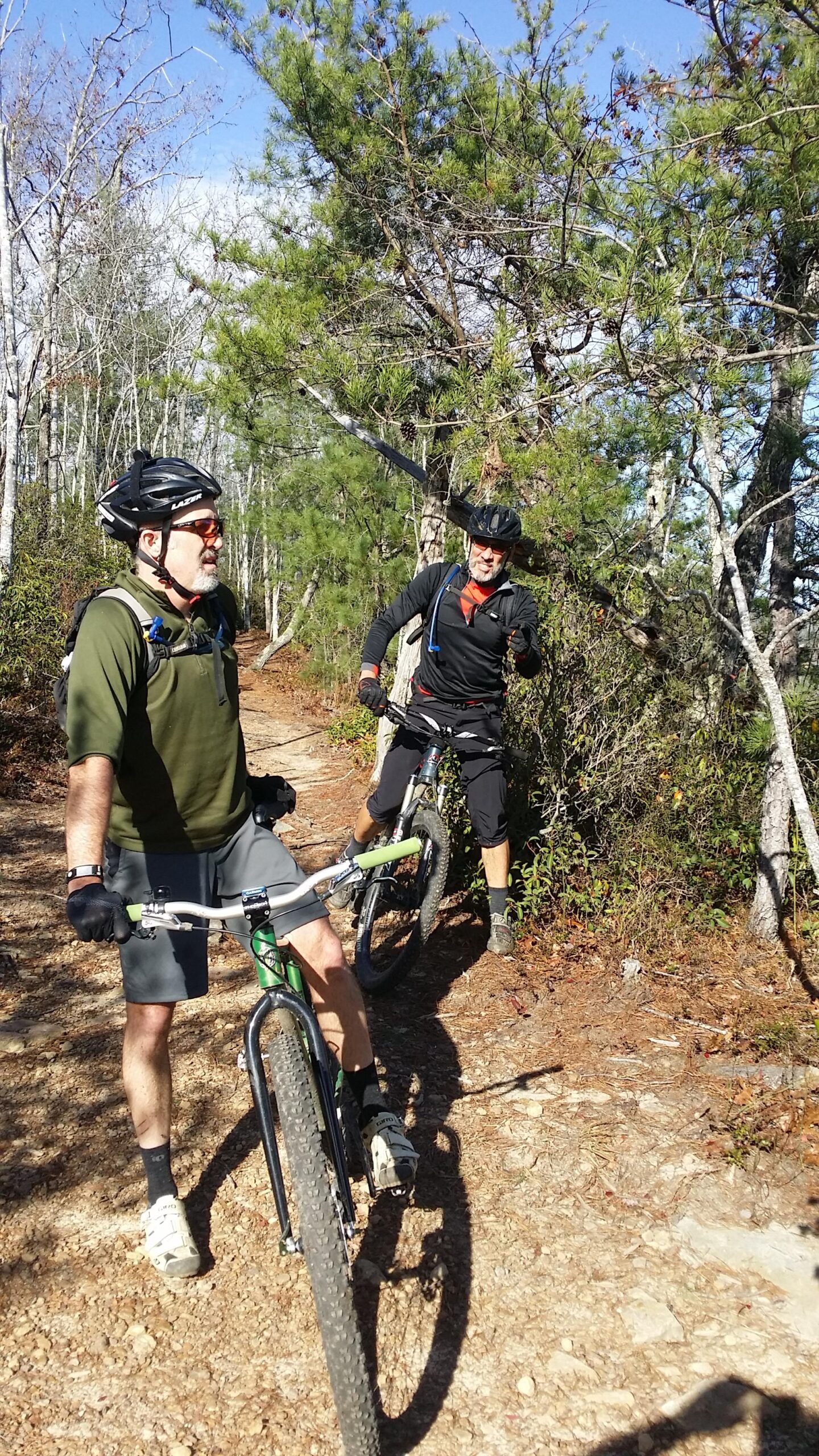 Two mountain bikers paused on a dirt trail surrounded by trees. One biker stands next to his green bike, wearing a green shirt and shorts, while the other, in a black outfit, gestures enthusiastically. The scene captures a sunny day in a natural setting, with clear blue skies and sparse foliage. Grand Gap mountain bike trail.