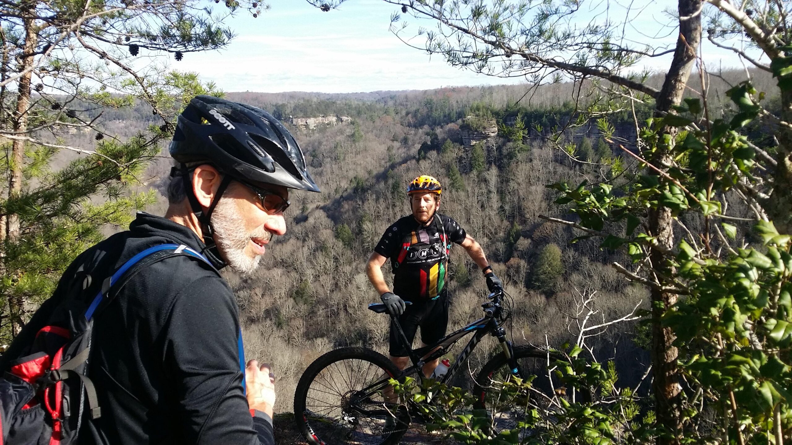 Two mountain bikers pause during their ride on a rocky trail overlooking a lush, wooded valley. One biker, wearing sunglasses and a helmet, is turned slightly to the side, while the second biker stands beside his bike, looking toward the camera. The background features a mix of trees and cliffs under a clear blue sky. Grand Gap mountain bike trail.