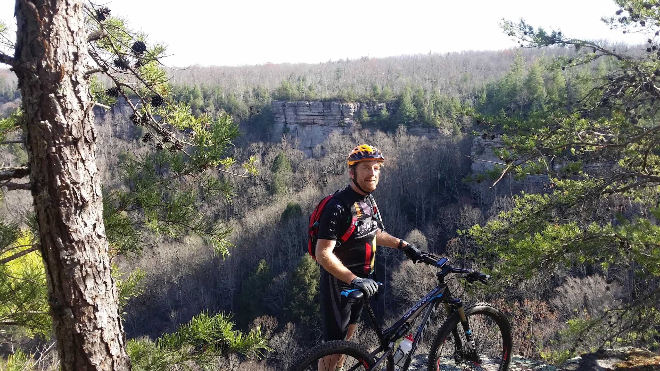 A person standing with a mountain bike on a rocky outcrop, overlooking a scenic view of a forested valley and cliffs. The individual is wearing a helmet and biking gear, surrounded by pine trees and clear blue skies. Grand Gap mountain bike trail.