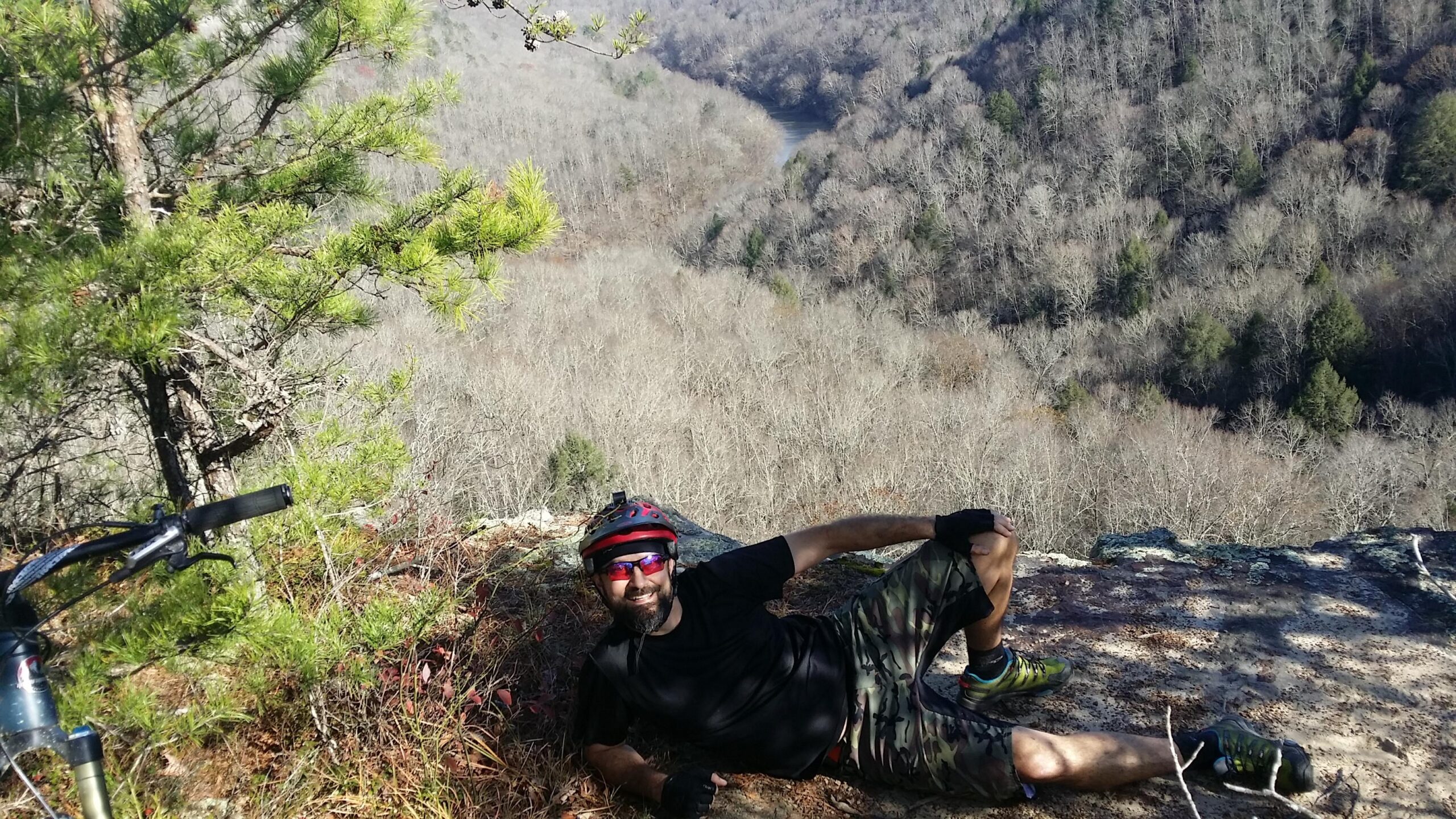 A mountain biker relaxing on a rocky overlook, wearing a helmet and sunglasses. The scene features a view of a river winding through a valley surrounded by bare trees and distant mountains under a clear sky. A bicycle is partially visible in the foreground, emphasizing the outdoor adventure theme. Grand Gap mountain bike trail.