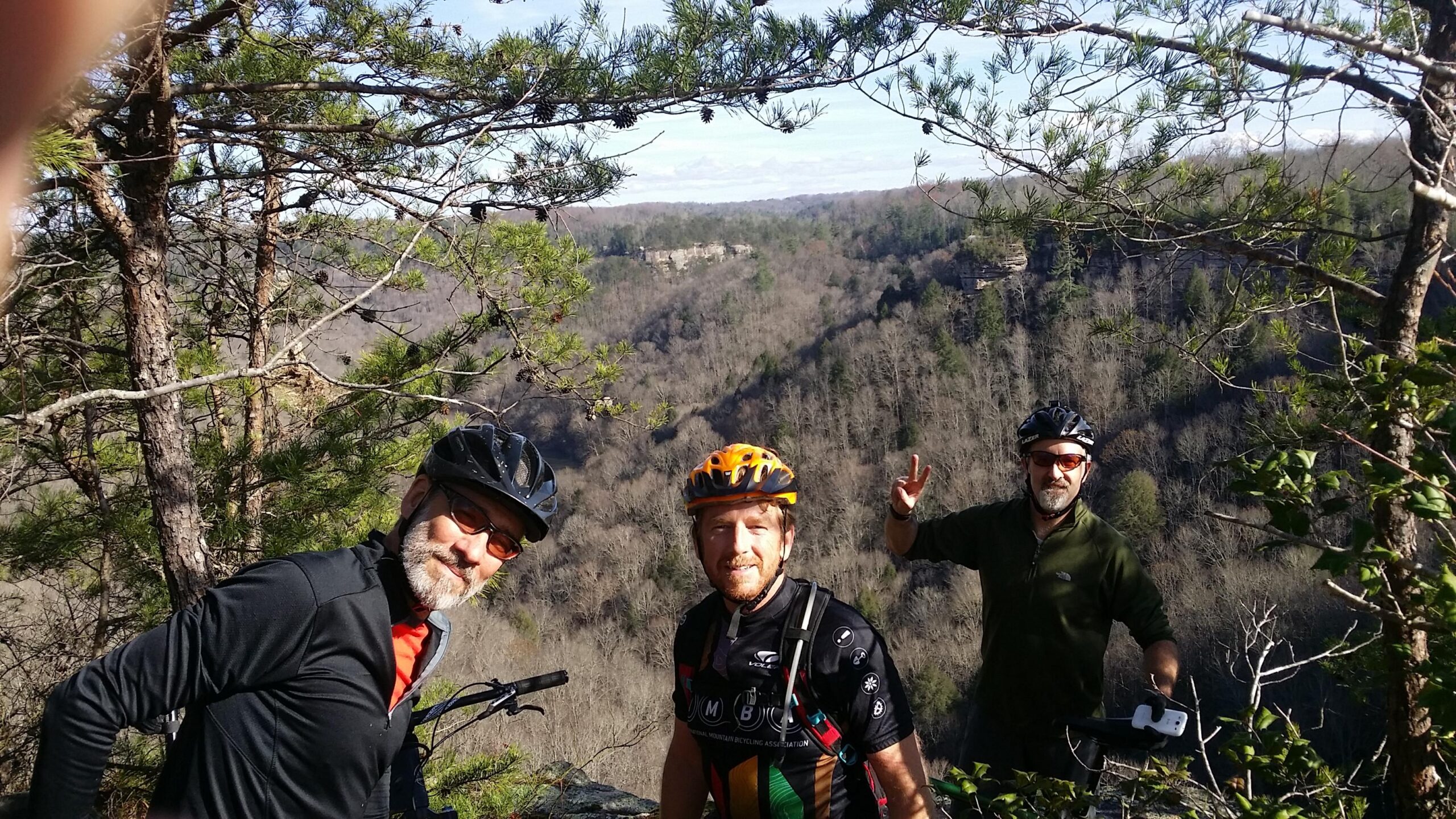 Three mountain bikers pose for a photo on a hillside, surrounded by trees and a scenic view of a valley below. The sun is shining, and the landscape features bare trees and distant hills. The first biker, in a black jersey and helmet, smiles at the camera, while the second, in an orange helmet, also grins. The third biker, in a green jacket and helmet, raises his hand in a peace sign. Grand Gap mountain bike trail.