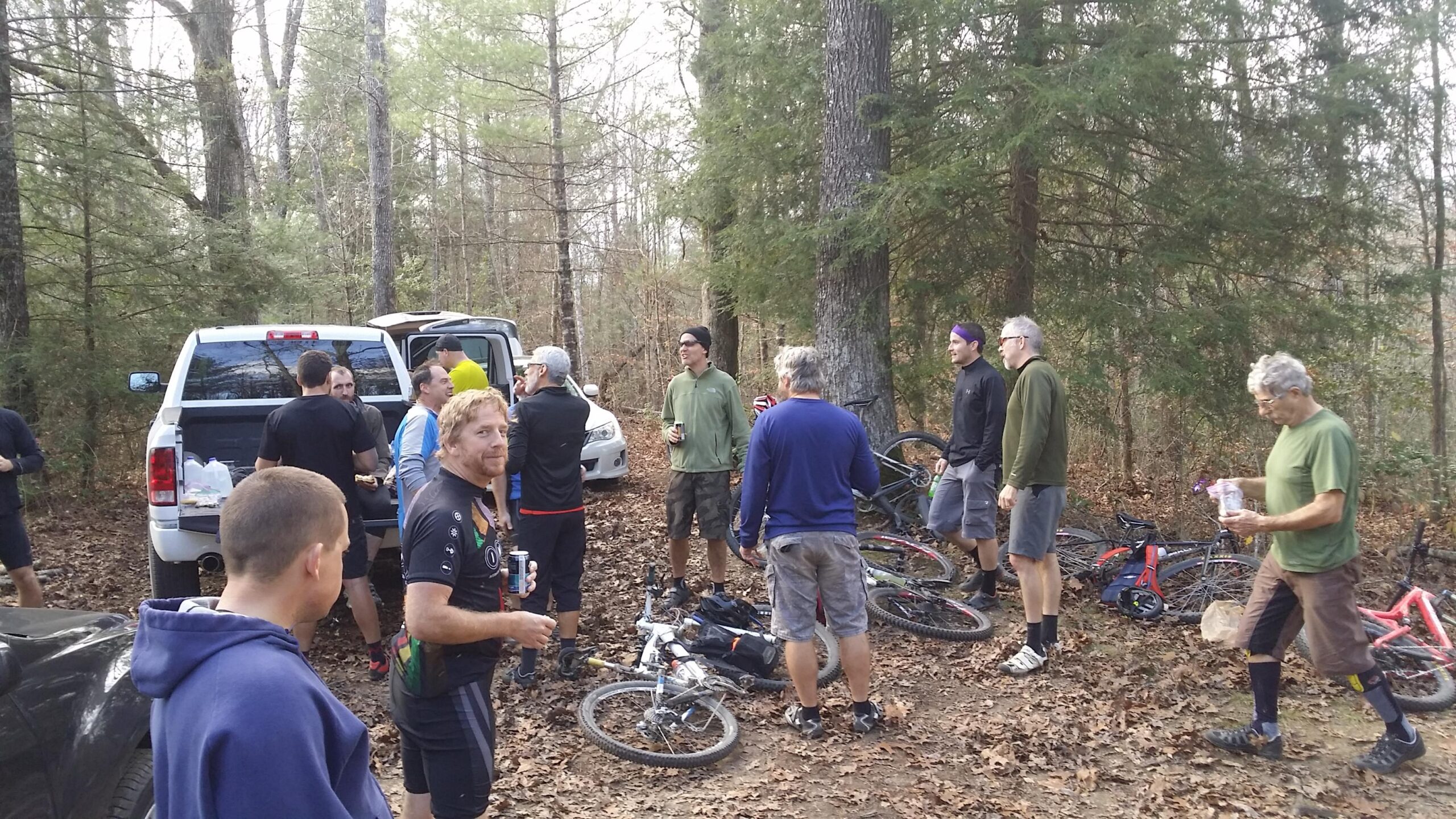 A group of mountain bikers gathered in a wooded area, socializing near a parked truck. Some individuals are standing and talking, while others are sitting or leaning against bicycles. The ground is covered in fallen leaves, and trees surround the scene. Several people are holding drinks and food as they enjoy a break after biking. Grand Gap mountain bike trail.