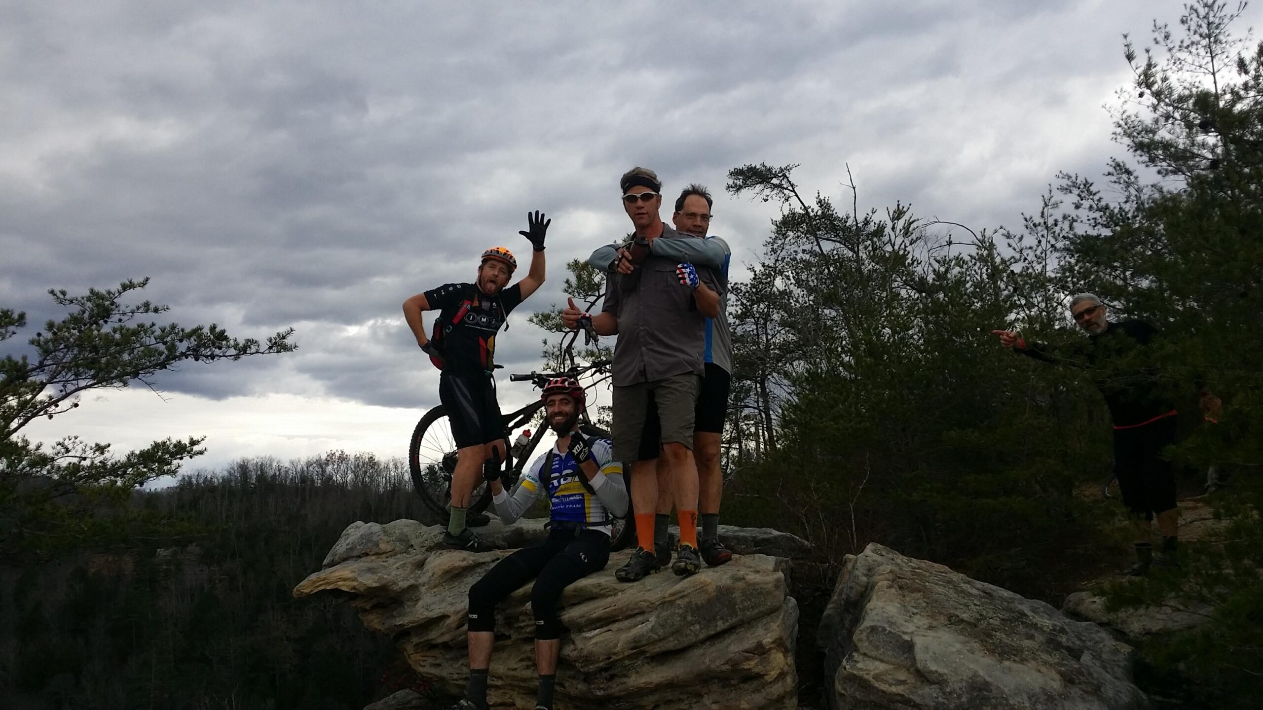 A group of five mountain bikers pose on a rocky outcrop surrounded by trees, with cloudy skies overhead. Two cyclists stand with their arms raised in excitement, while another sits on the rock below, flashing a peace sign. A fourth cyclist is behind them, and a fifth rider is partially visible in the background, pointing towards the camera. Grand Gap mountain bike trail.
