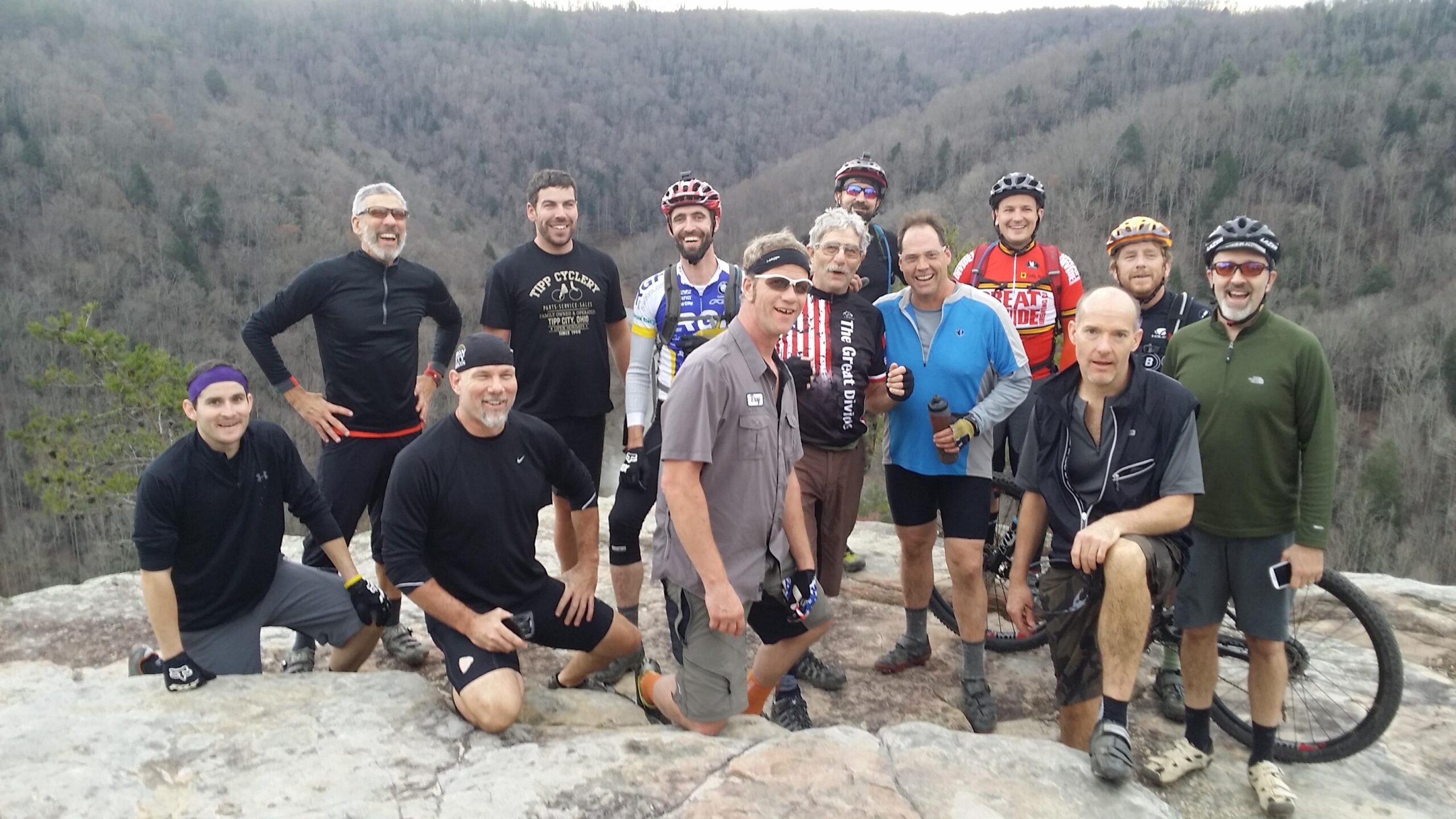 A group of thirteen mountain bikers posing together on a rocky outcrop with a scenic forested landscape in the background. They are smiling and wearing cycling gear, with some holding water bottles and bikes. The atmosphere is joyful and energetic, suggesting a sense of camaraderie and outdoor adventure. Grand Gap mountain bike trail.