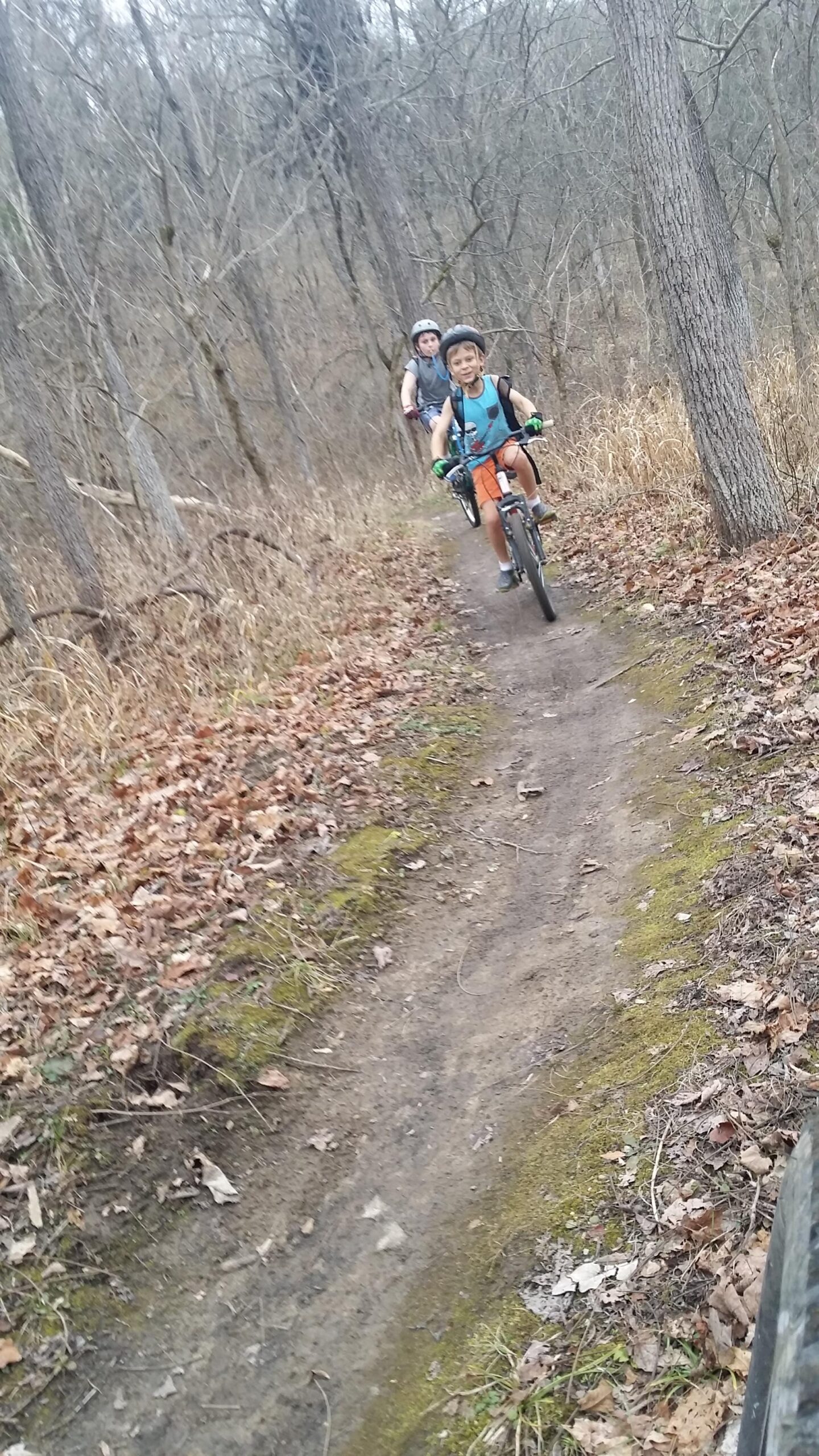 Two children riding bicycles on a dirt trail through a wooded area. The child in the foreground is smiling and wearing a helmet, while the other child follows behind, also wearing a helmet. The trail is surrounded by bare trees and fallen leaves, indicating an early spring or late fall setting. Versailles State Park mountain bike trail.
