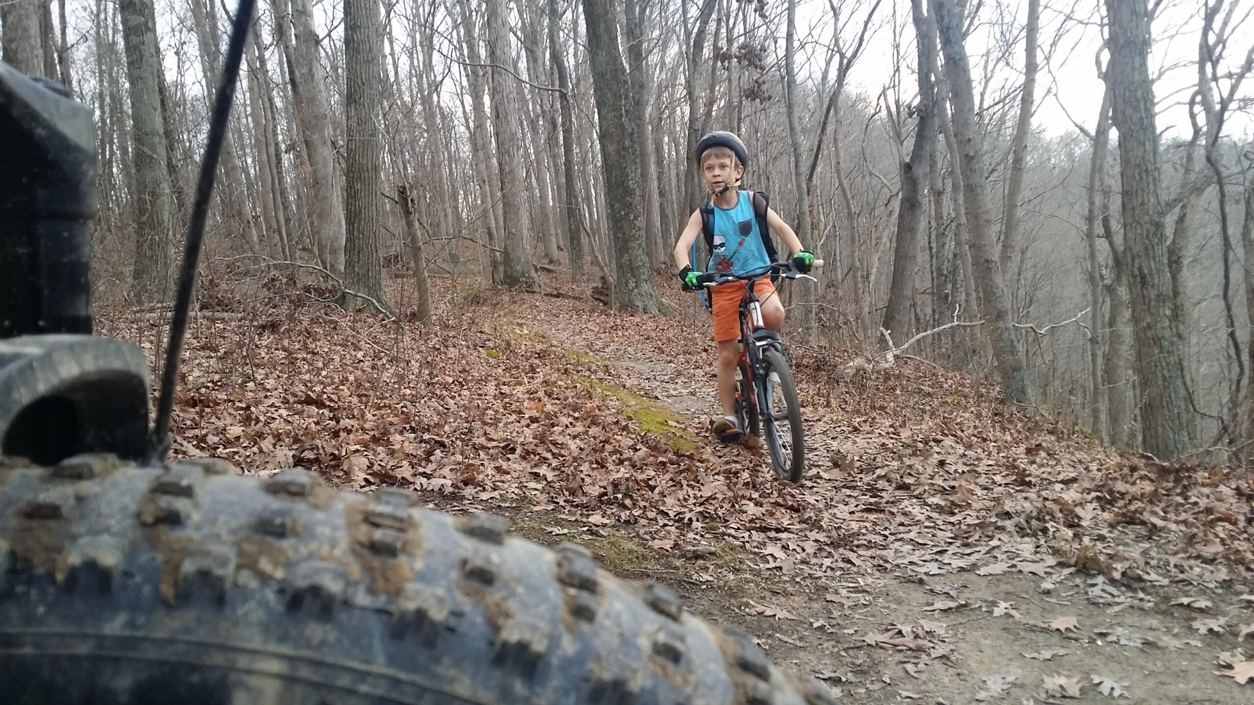 A young boy riding a bicycle along a wooded trail, surrounded by bare trees and fallen leaves. He is wearing a tank top, shorts, and a helmet, with a backpack and green gloves. In the foreground, part of a bicycle tire is visible. Versailles State Park mountain bike trail.