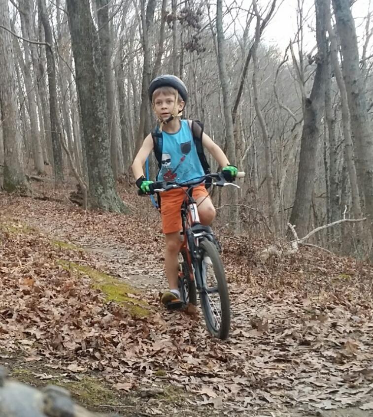 A young boy wearing a helmet and colorful athletic gear rides a mountain bike along a leaf-covered trail in a wooded area, surrounded by bare trees. Versailles State Park mountain bike trail.