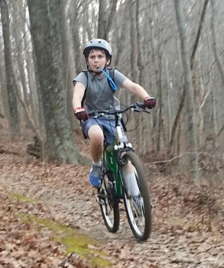 A young boy riding a green mountain bike along a wooded trail, wearing a helmet and casual clothing. He appears focused and is navigating over a leaf-covered path surrounded by trees. Versailles State Park mountain bike trail.