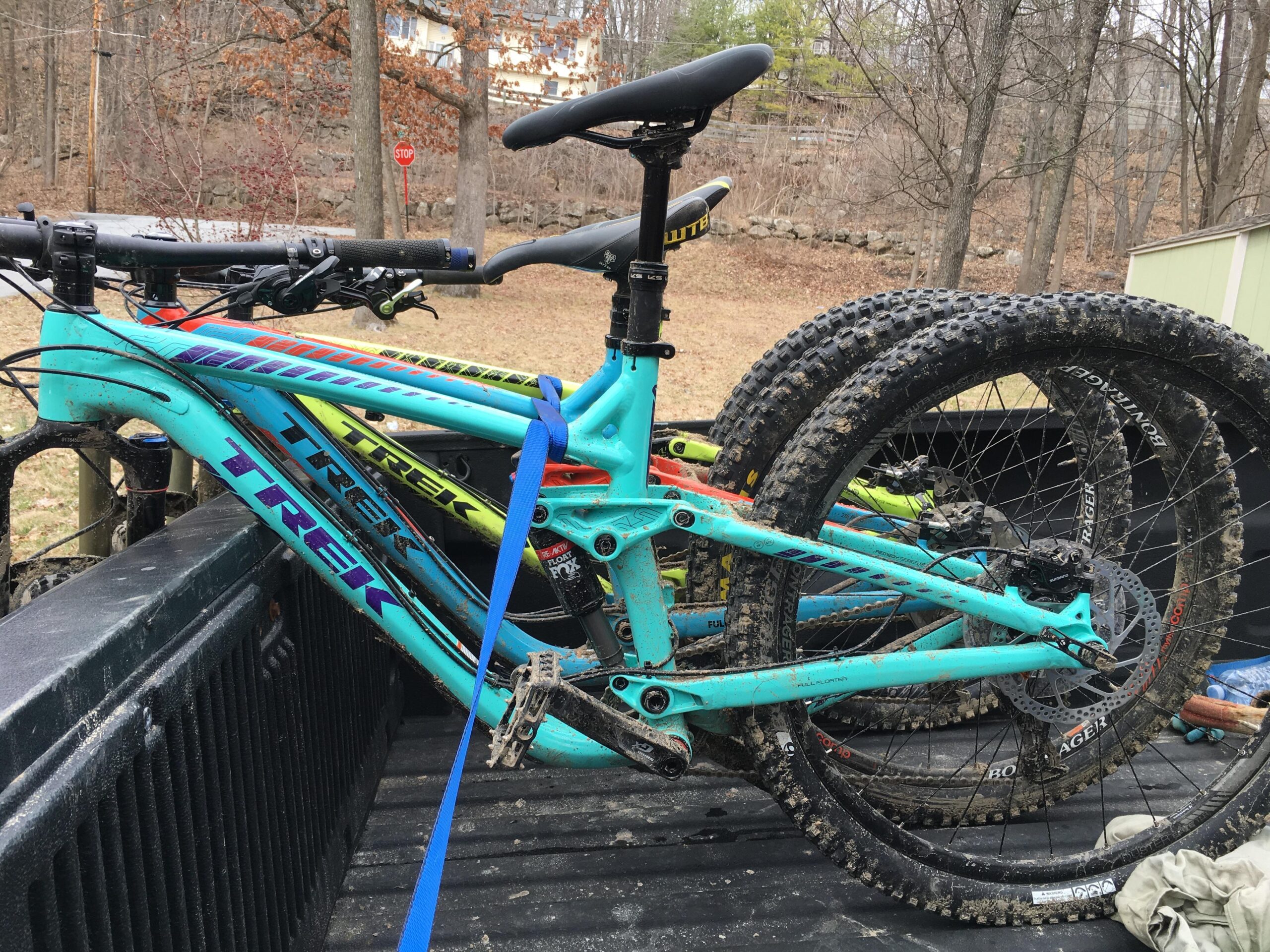 Trek Remedy 8 27.5/650b: Two mountain bikes with a turquoise frame are secured on the back of a pickup truck. The bikes show signs of mud and wear, indicating recent use on a trail. One bike is positioned upright, while the other is leaned against it, with their tires partially visible. The background features bare trees and a glimpse of a house, along with a stop sign.