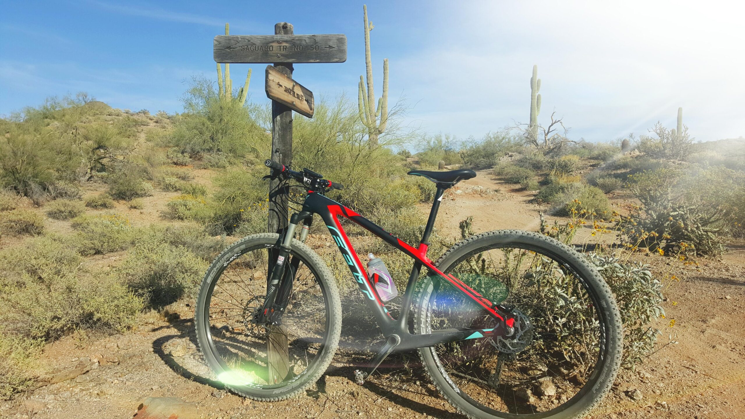 A mountain bike parked next to a trail sign in a desert landscape with cacti and shrubs. The sign indicates "Saguaro Trail No. 50," surrounded by rocky terrain and under a blue sky. Hawes Loop mountain bike trail.