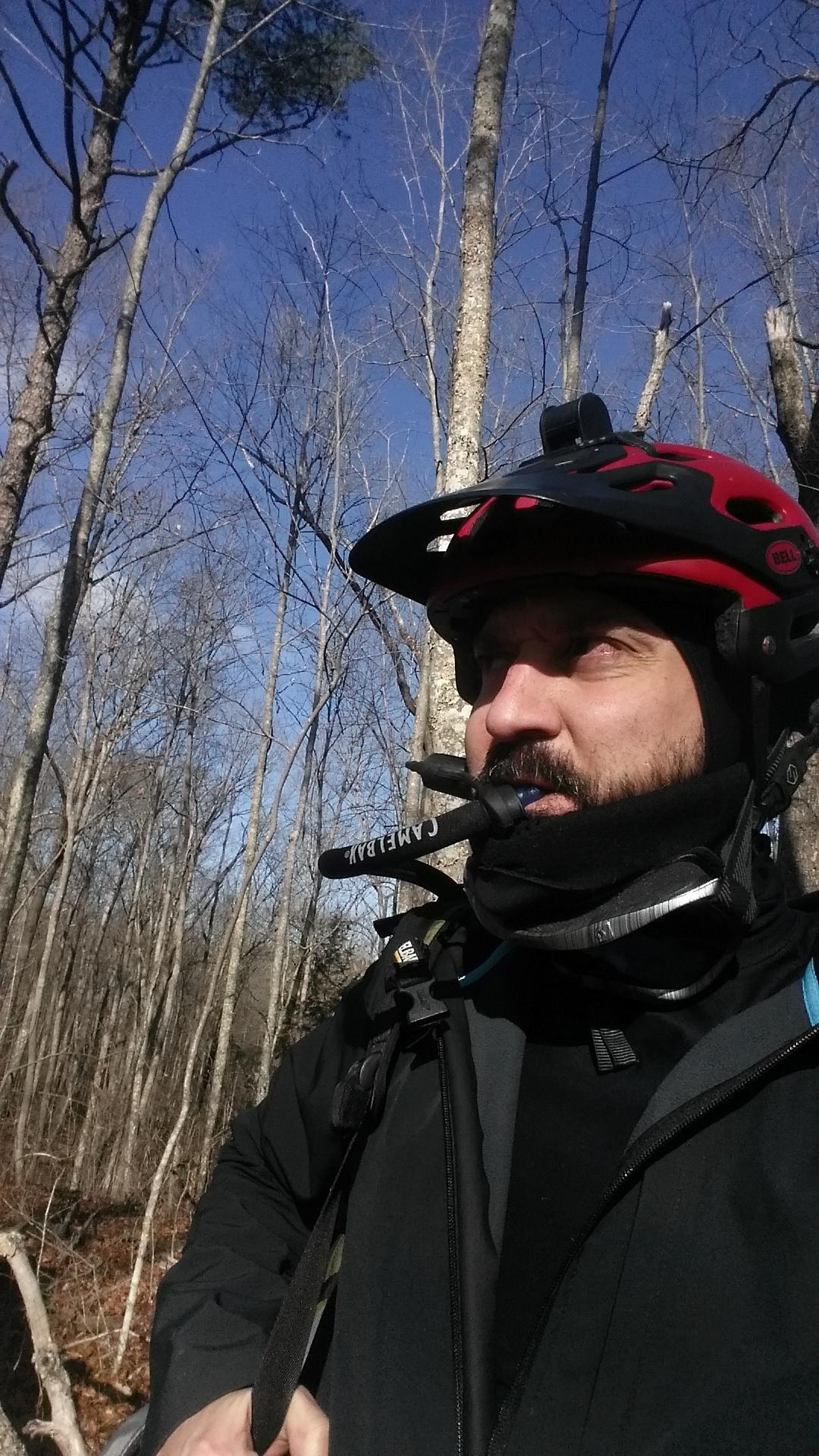 A person wearing a red and black helmet and a black jacket, standing outdoors in a wooded area. The background features tall, bare trees against a blue sky. The individual appears to be using a hands-free device, with a water tube visible near their mouth. Versailles State Park mountain bike trail.