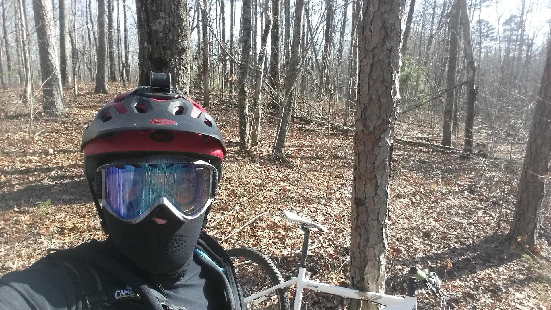 A person wearing a black mask and helmet with a red and black design stands in a wooded area. They are holding a mountain bike with a white frame beside them. The forest floor is covered with leaves, and trees surround the scene in bright daylight. Versailles State Park mountain bike trail.