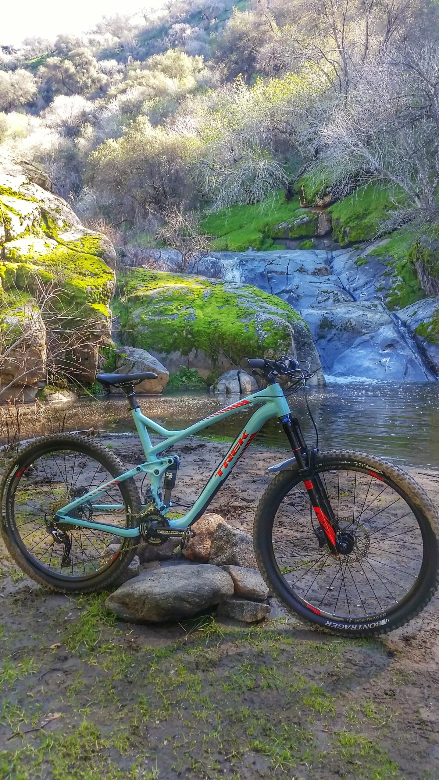 Trek trek remedy 7 27.5: A mountain bike resting on rocks near a small stream and waterfall, surrounded by lush greenery and moss-covered boulders in a natural outdoor setting.