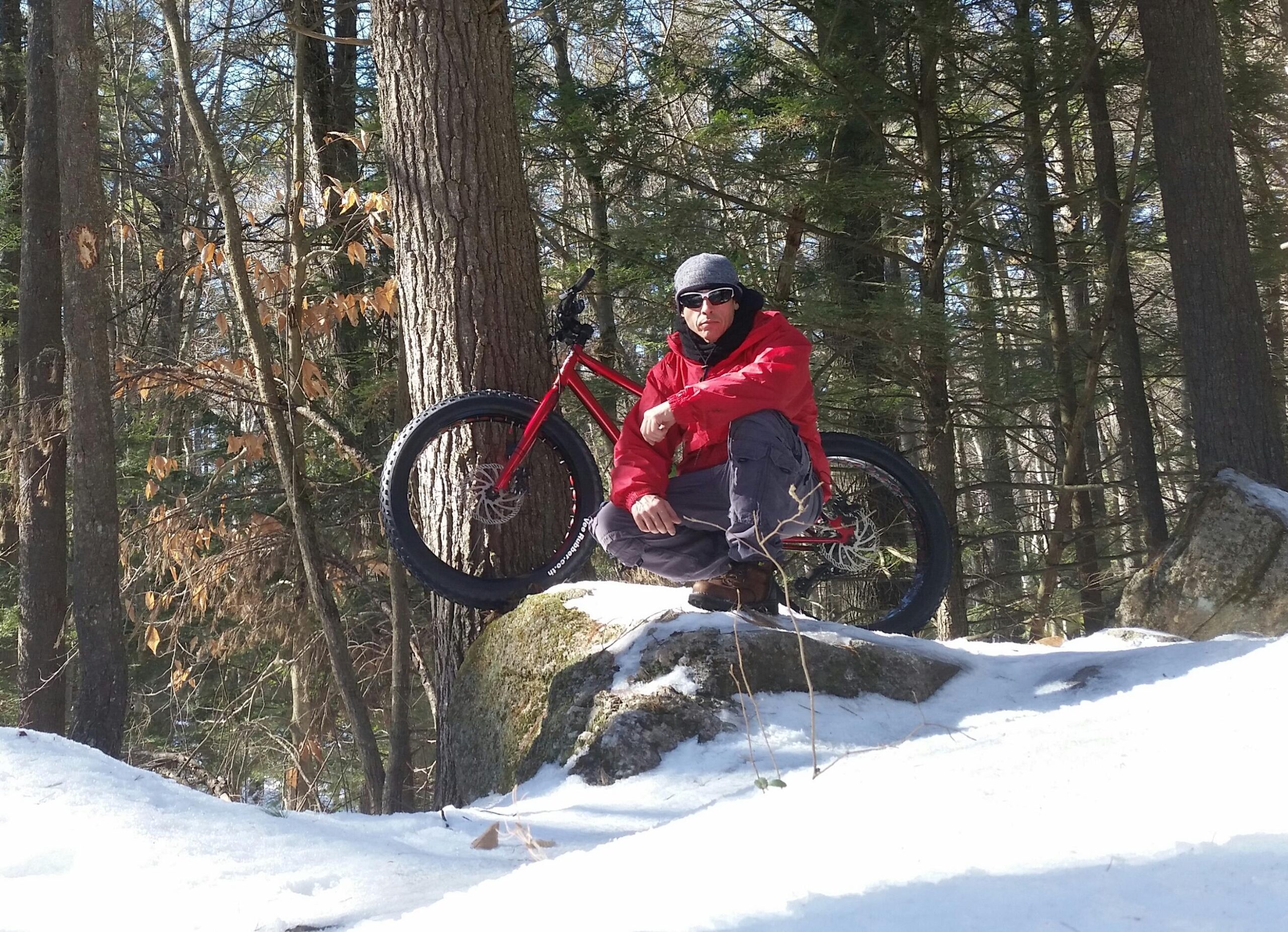 A person in a red jacket and gray pants kneels on a rock covered in snow, beside a red fat bike with large tires. The background features a snow-covered forest with tall trees and some remaining autumn leaves. Depot Road Singletracks/FOMBA mountain bike trail.