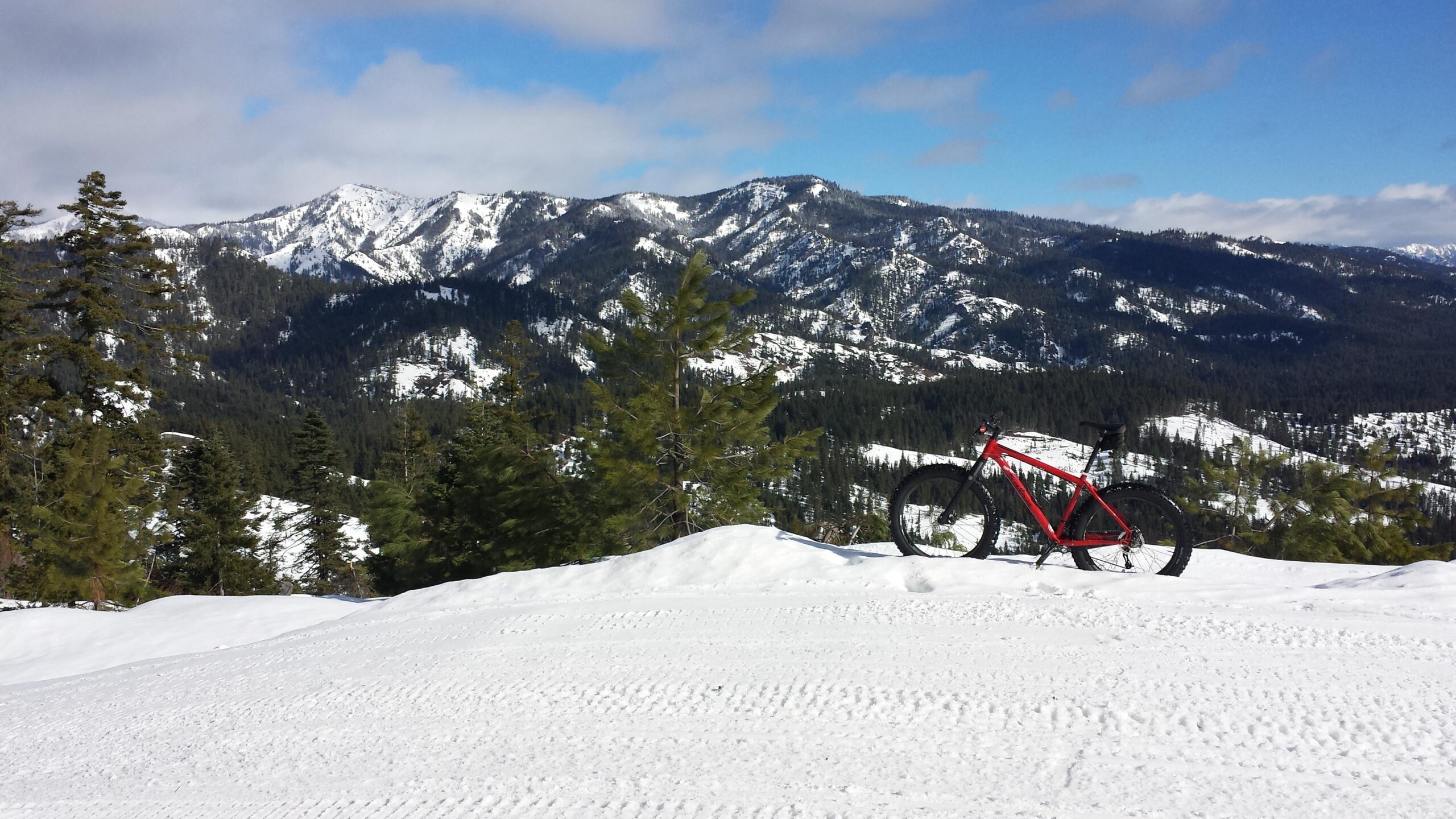 Salsa Mukluk: A red fat tire bike resting on a snow-covered landscape, with a backdrop of snow-capped mountains and evergreen trees under a partly cloudy blue sky.