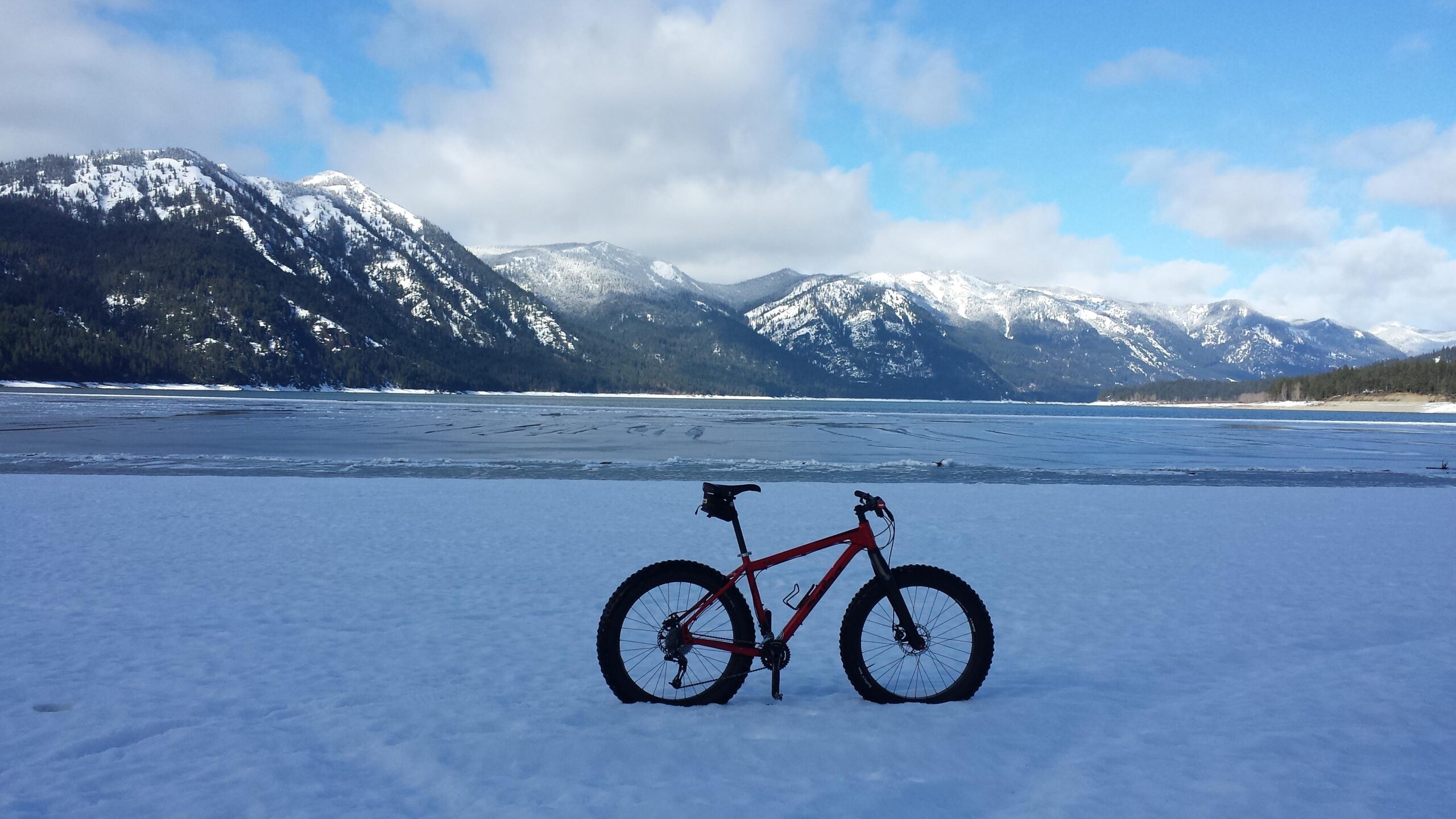Salsa Mukluk: A red fat bike stands on a snowy landscape with a frozen lake in the background, framed by snow-capped mountains under a partly cloudy blue sky.