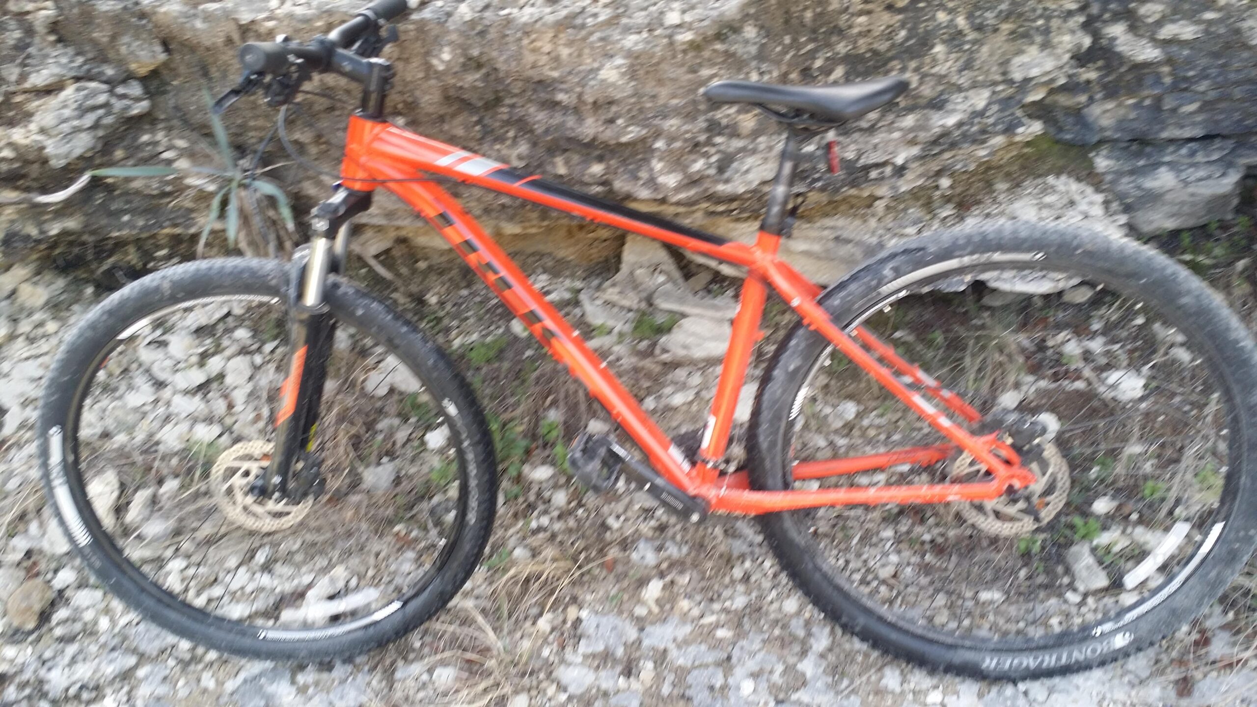 Trek X-Caliber 6: A close-up view of an orange mountain bike resting against a rocky surface. The bike features thick tires suitable for off-road terrain, and a simple black seat. The surrounding area includes gravel and some sparse vegetation.