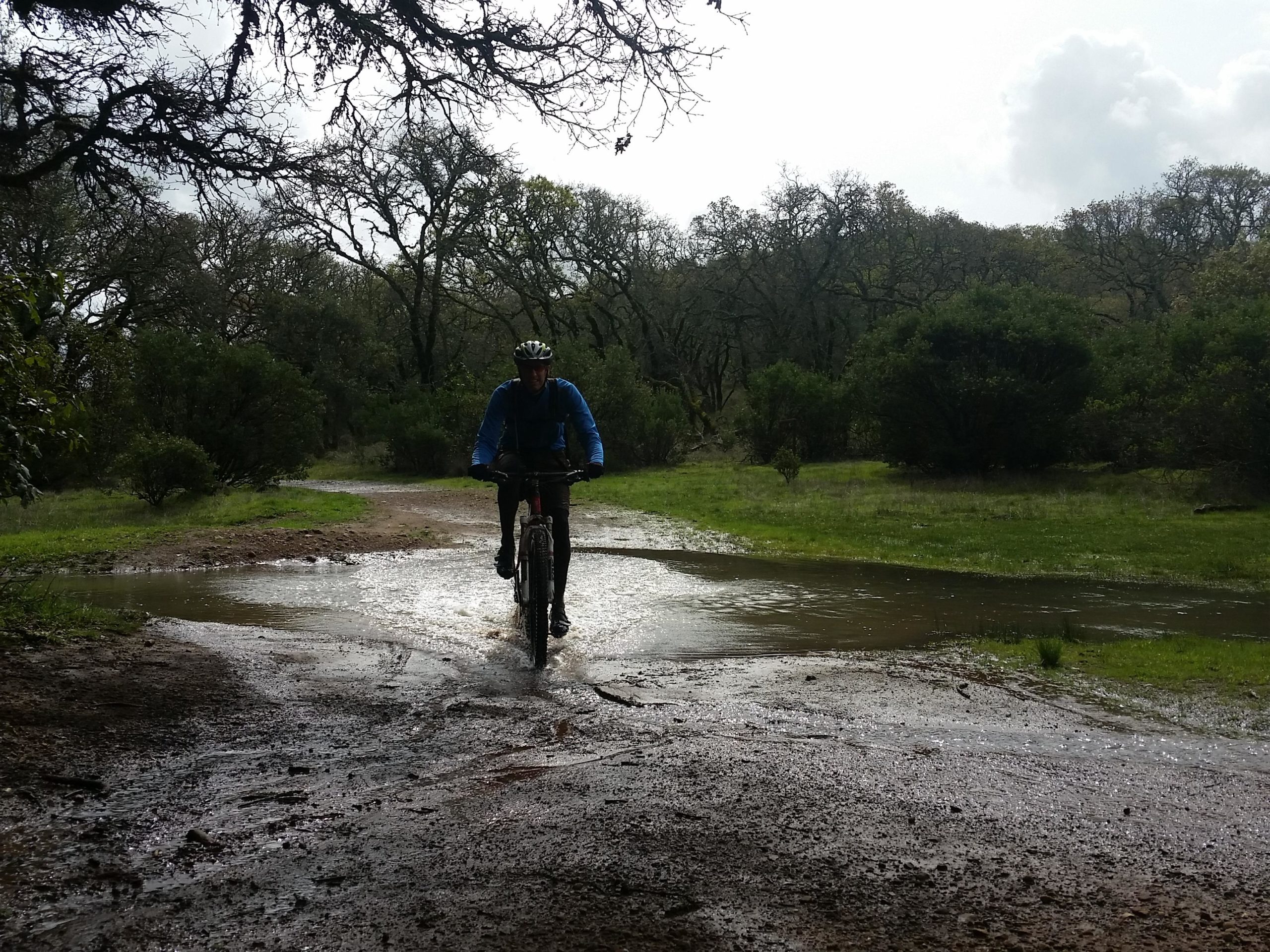 A mountain biker rides through a muddy trail with a small water crossing, surrounded by greenery and trees. The sky is partly cloudy, suggesting a recent rain. Annadel State Park mountain bike trail.