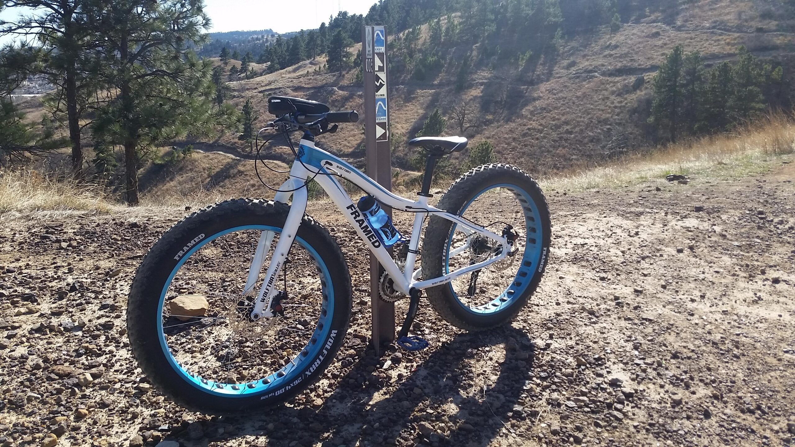 Framed Framed Wolftrax: A white fat tire mountain bike with blue accents parked beside a trail marker in a rugged outdoor landscape featuring rolling hills and pine trees under a clear sky.