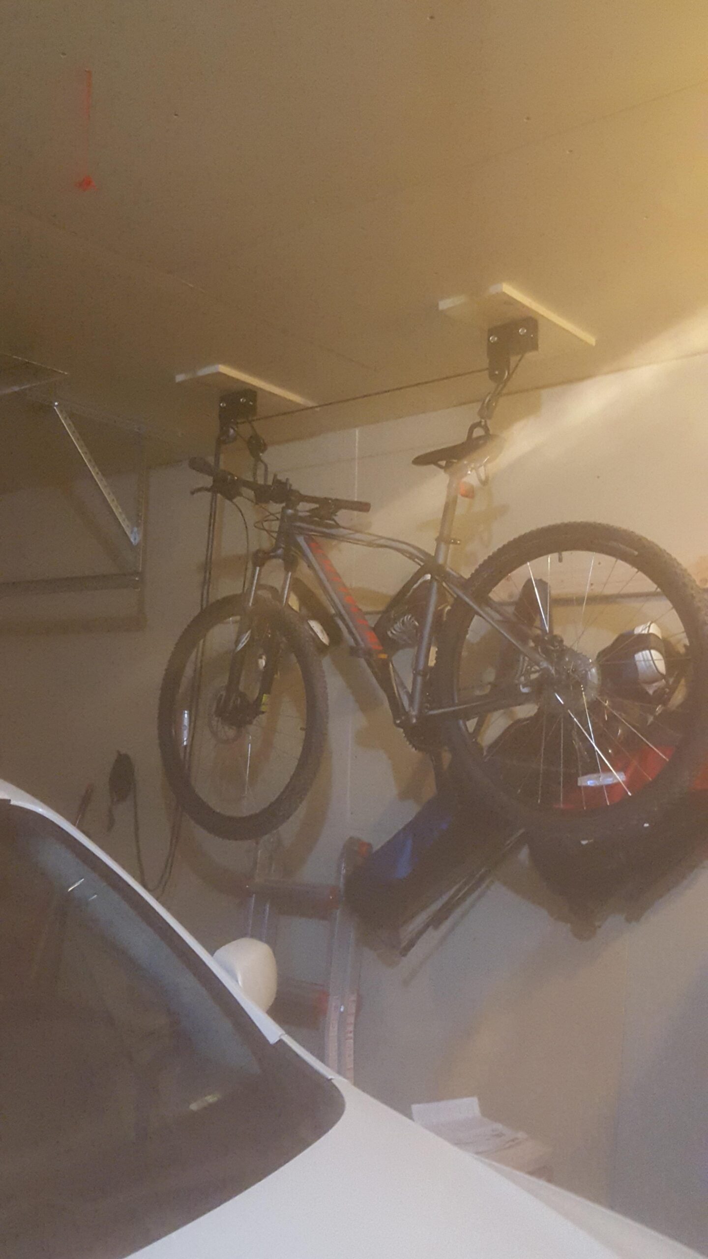 Specialized Rockhopper: A mountain bike suspended from the ceiling in a garage, with a white car partially visible in the foreground. The garage includes shelves and various items stored nearby, creating a cluttered but organized space. The bike is secured with ceiling-mounted hooks.