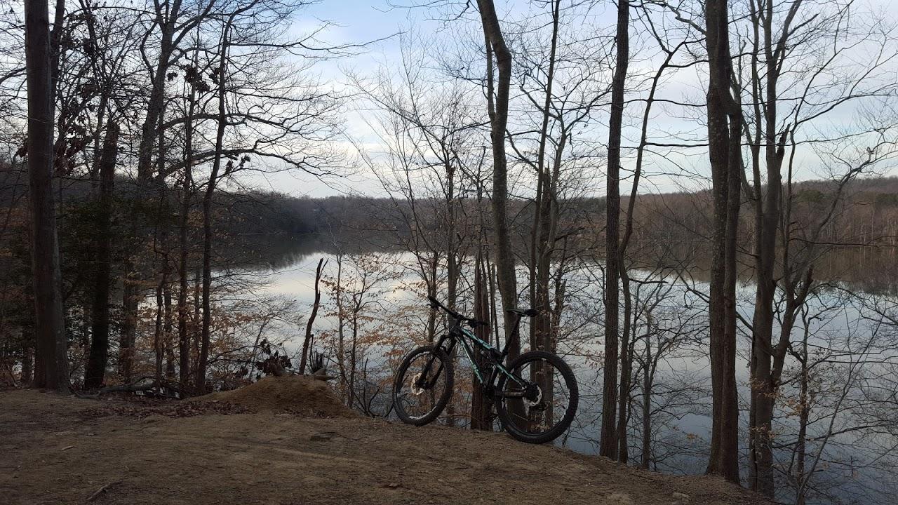 A mountain bike parked on a dirt trail overlooking a calm lake surrounded by bare trees, with a cloudy sky reflecting in the water. Fountainhead Regional Park mountain bike trail.