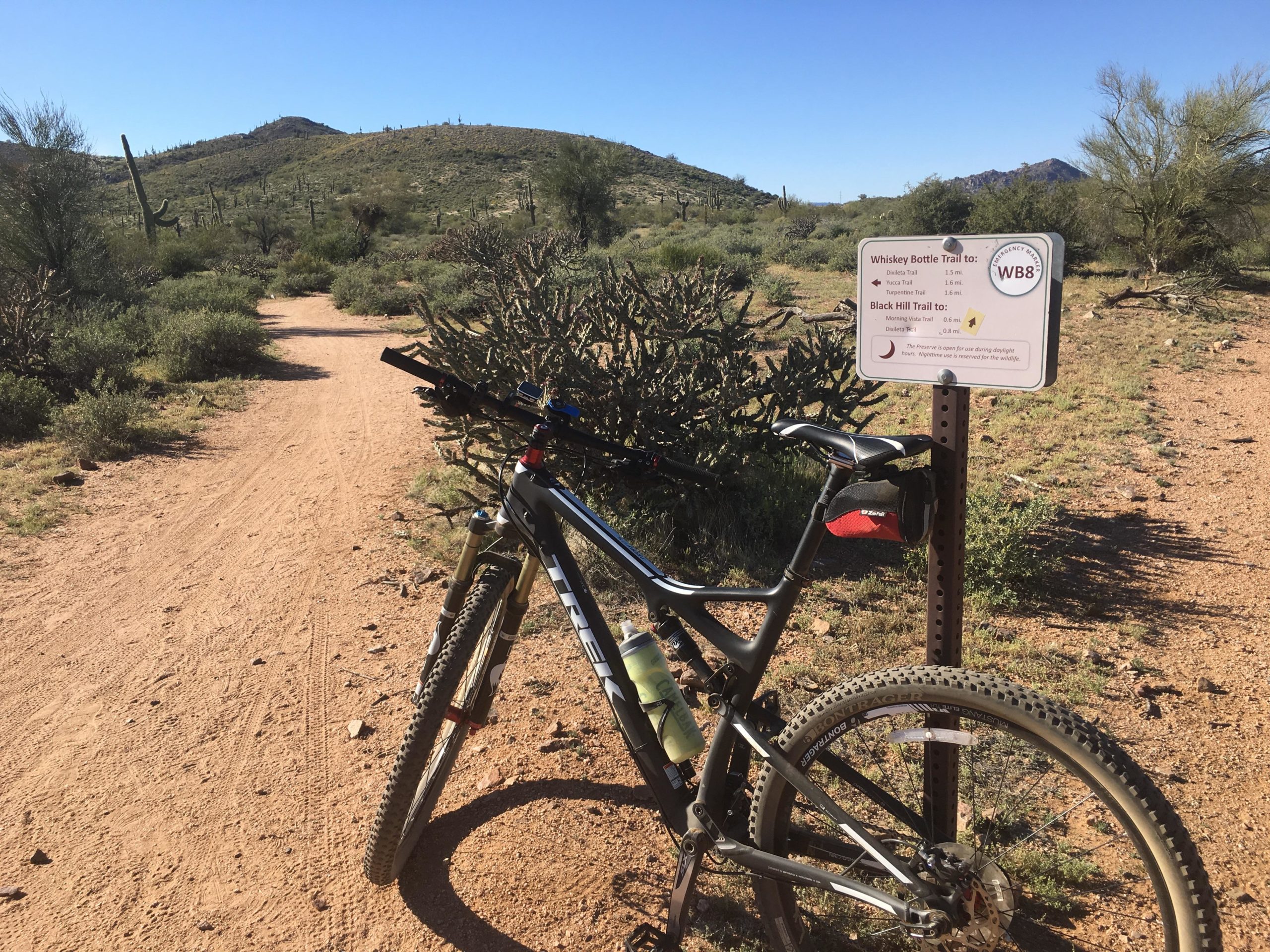 A mountain bike parked next to a trail sign indicating directions for Whiskey Bottle Trail and Black Hill Trail. The trail is dirt with a surrounding landscape of desert vegetation, including cacti and shrubs, against a backdrop of rolling hills under a clear blue sky. Brown's Ranch to Granite Mountain mountain bike trail.