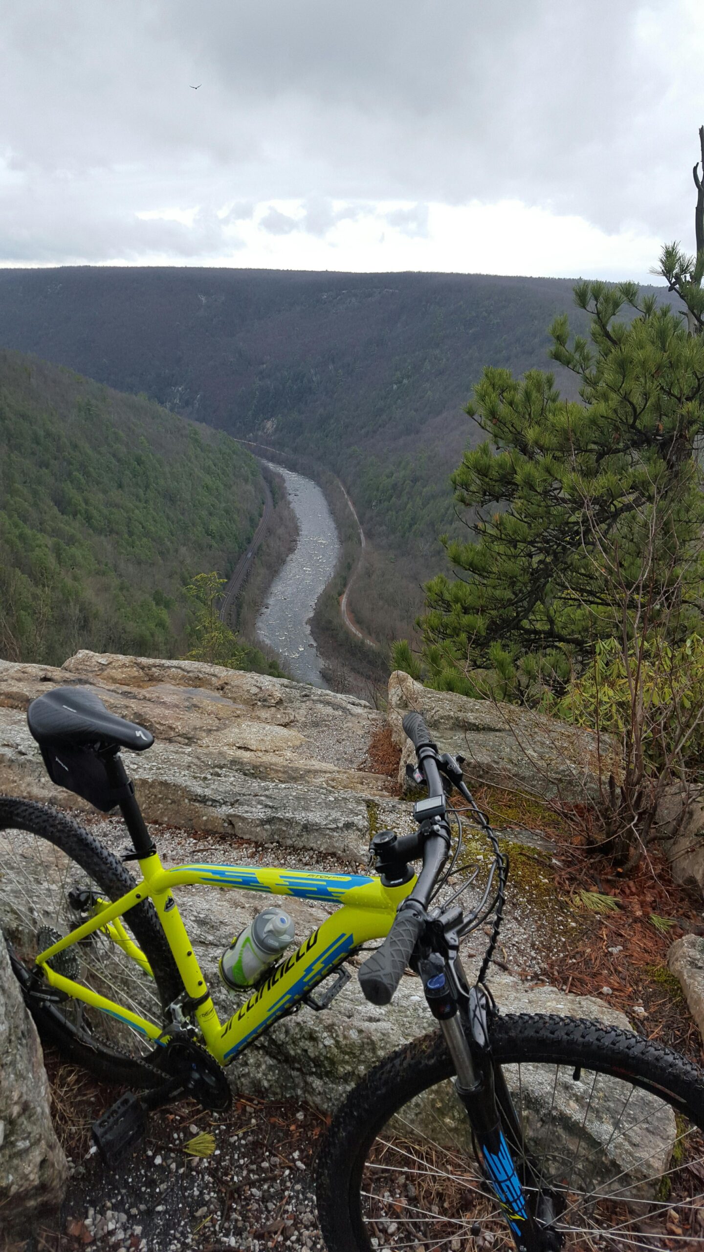 Specialized Pitch 650b: A vibrant yellow mountain bike is positioned on a rocky ledge overlooking a winding river nestled between green hills and valleys under a cloudy sky. The bike has a water bottle attached, and the scene conveys a sense of adventure and the beauty of nature.