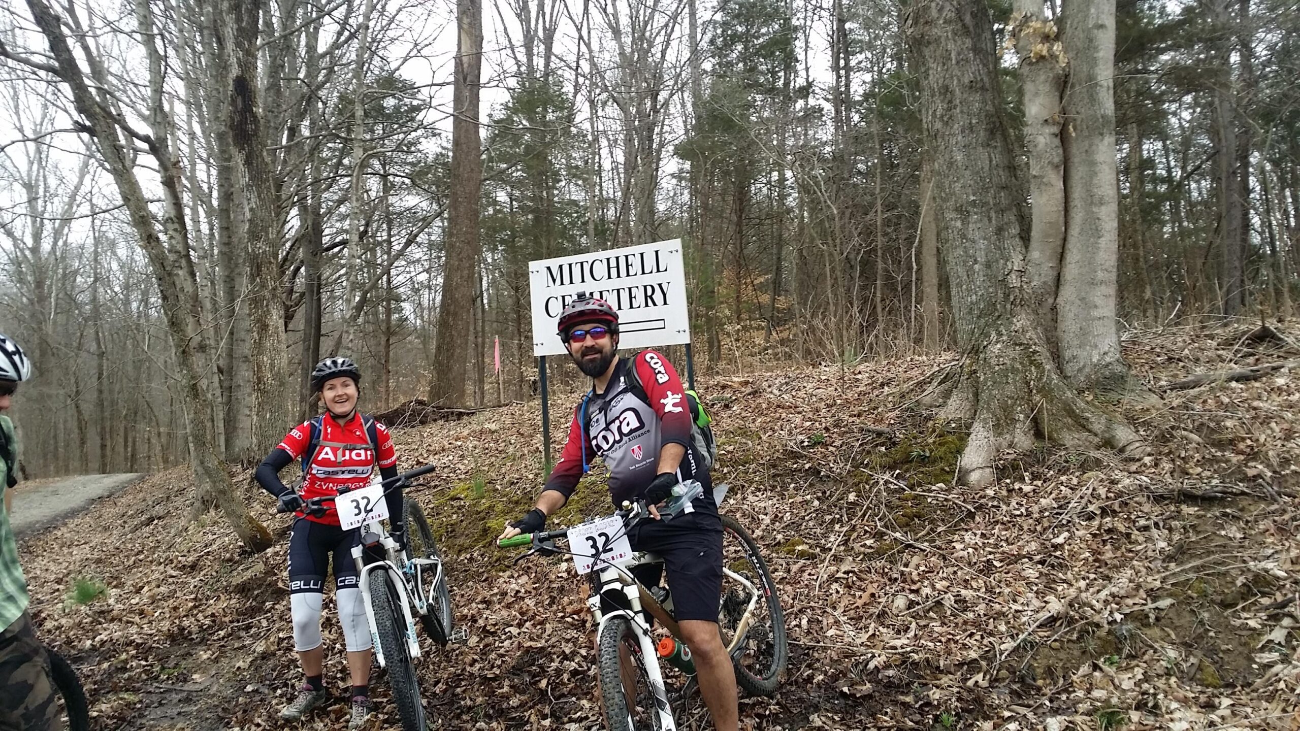 Two mountain bikers pose in front of a sign that reads "Mitchell Cemetery" in a wooded area. The male biker, wearing a black and red jersey, is seated on his bike with a friendly smile. The female biker stands next to him, also smiling, wearing a red jersey and white pants. The background features leafless trees and a path partially covered with leaves. Hickory Ridge mountain bike trail.