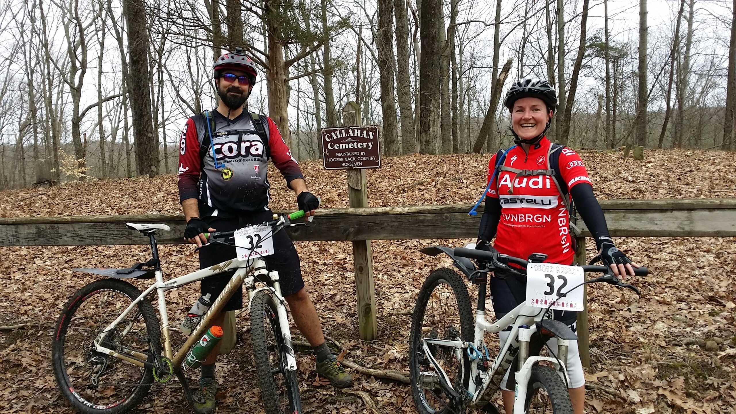 Two mountain bikers pose next to a sign for Callahan Cemetery in a forested area. The man on the left is wearing a black and red cycling jersey with the logo "Cora" and is holding a green water bottle, while the woman on the right wears a red jersey with "Audi" written on it. Both are standing beside their bikes and are surrounded by fallen leaves and tall trees, indicating a trail setting. Hickory Ridge mountain bike trail.