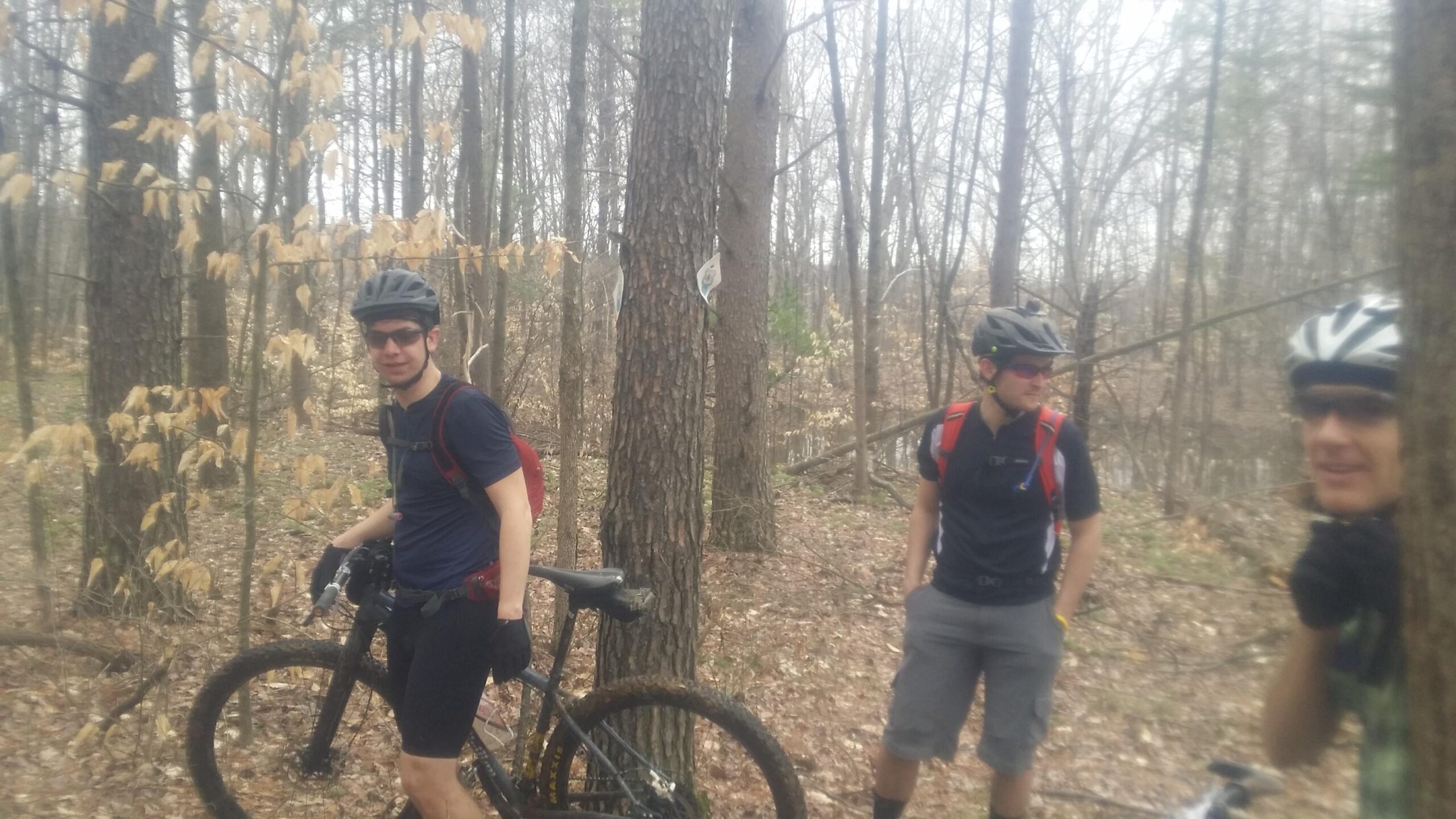 Three mountain bikers pause in a wooded area, surrounded by bare trees and fallen leaves. One rider, wearing a helmet and black sunglasses, stands next to his bike while the others chat nearby, dressed in cycling attire. The atmosphere suggests a break during an outdoor biking adventure. Hickory Ridge mountain bike trail.