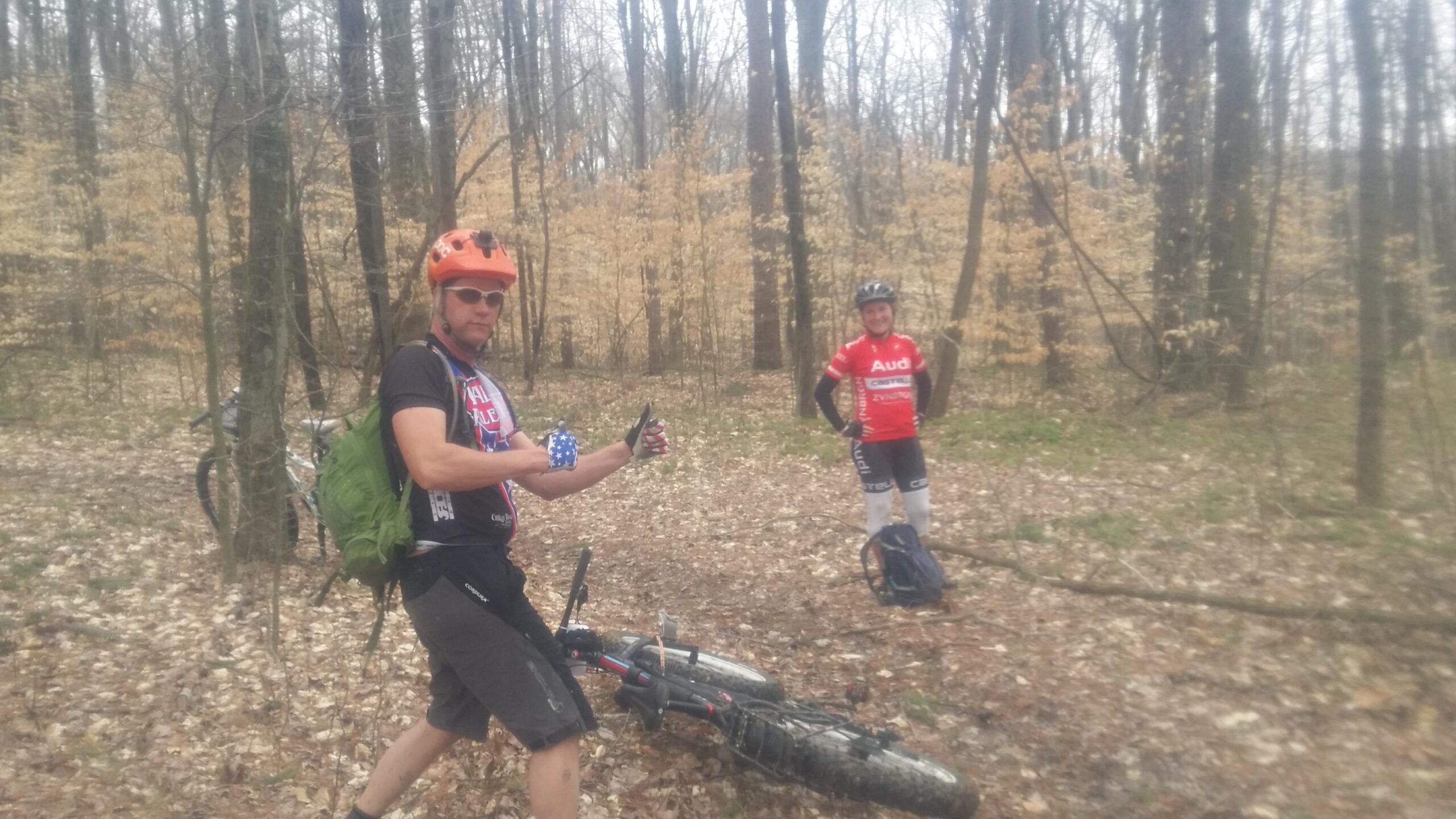 Two mountain bikers posing in a wooded area during springtime. One biker, wearing an orange helmet and a black shirt with an American flag design, is holding up a drink and smiling. The other biker, dressed in a red jersey, is standing nearby with their bike on the ground. The background features bare trees and fallen leaves. Hickory Ridge mountain bike trail.